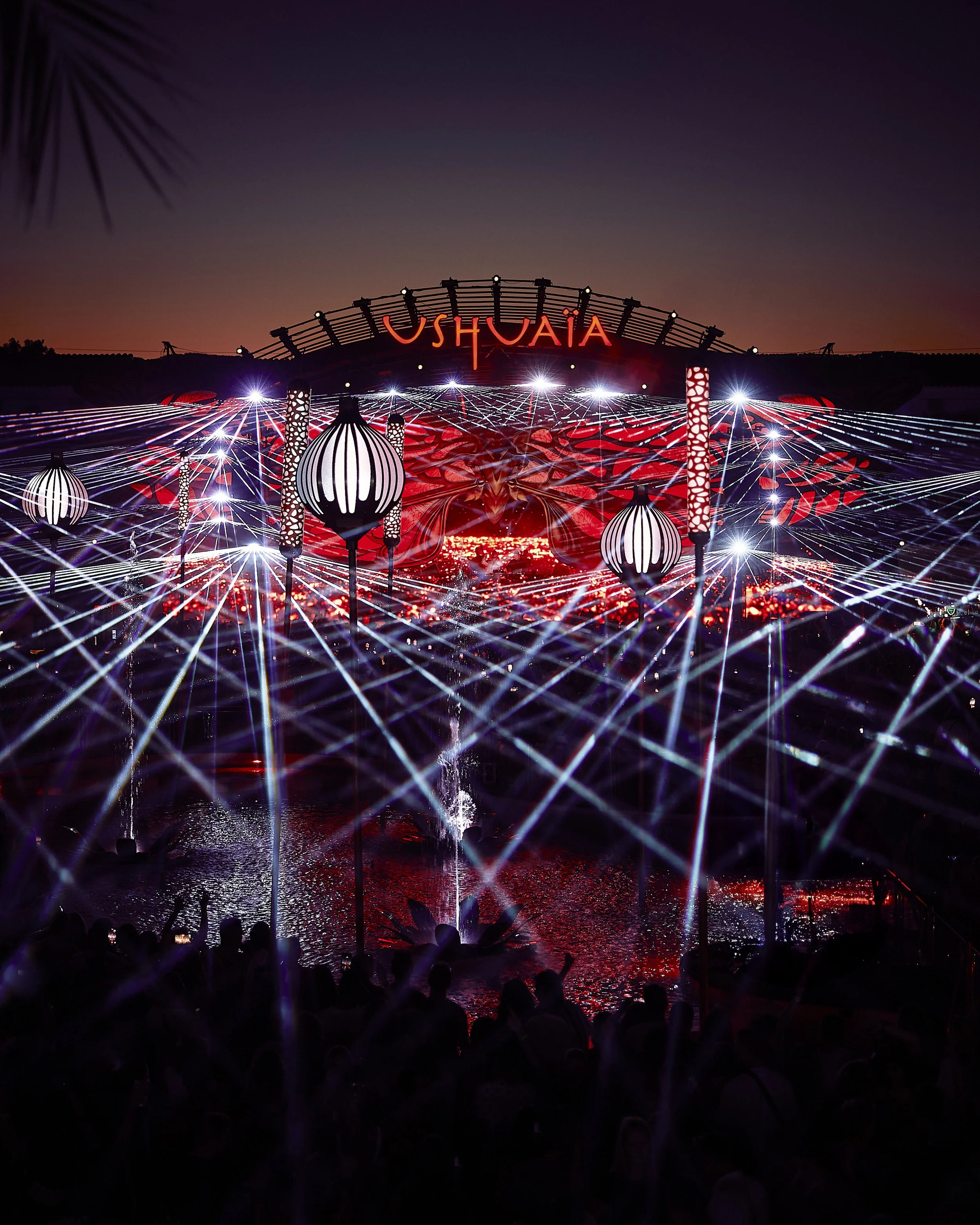 A vibrant nighttime scene at a music festival with colorful lights, laser beams, and large lantern-like structures, with the audience visible in silhouette in the foreground and the word 'USHUAÏA' illuminated in the background.