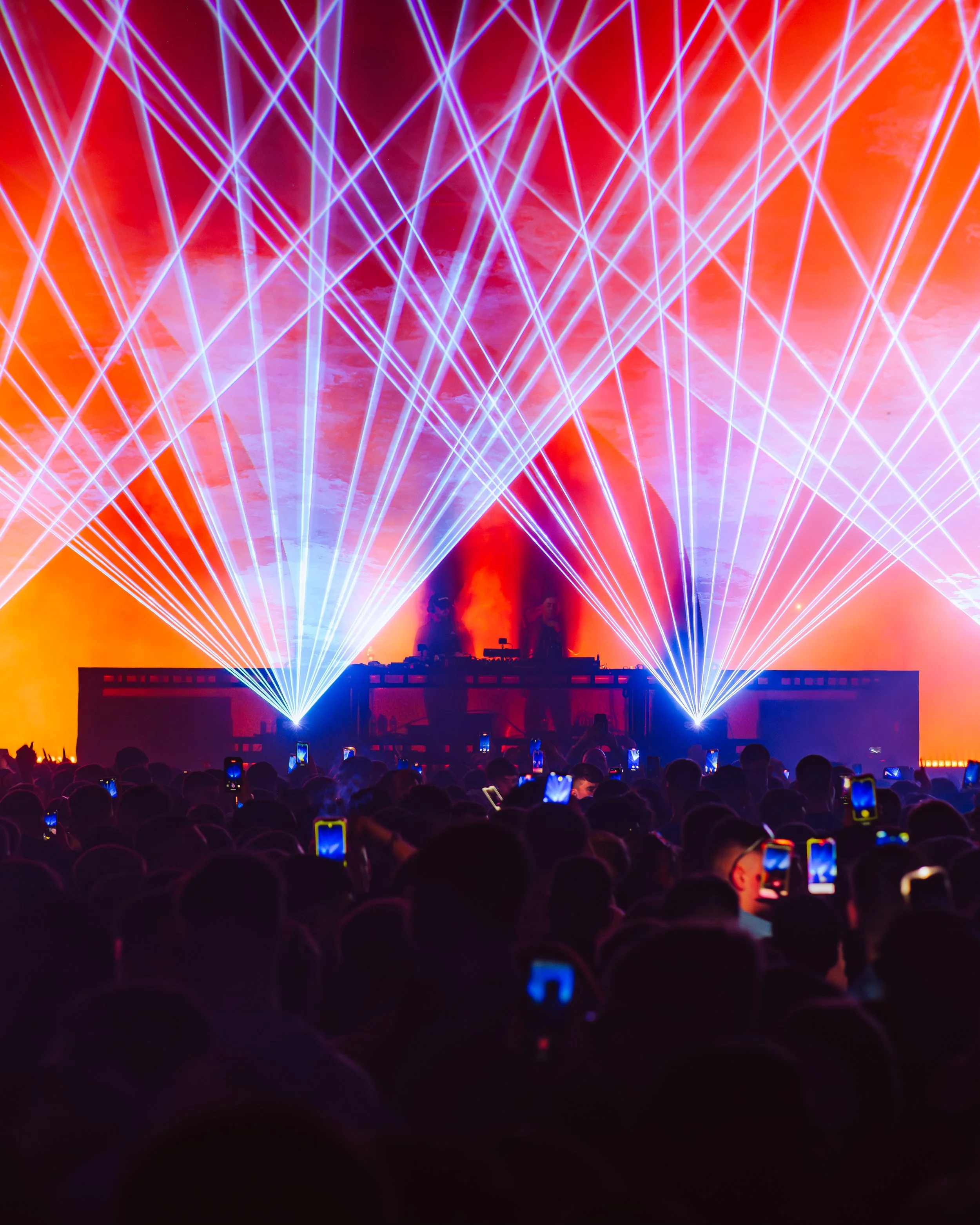 A large crowd watching an outdoor laser light show with bright beams of blue, purple, and orange lights shooting into the sky, and silhouettes of people holding smartphones in the foreground.
