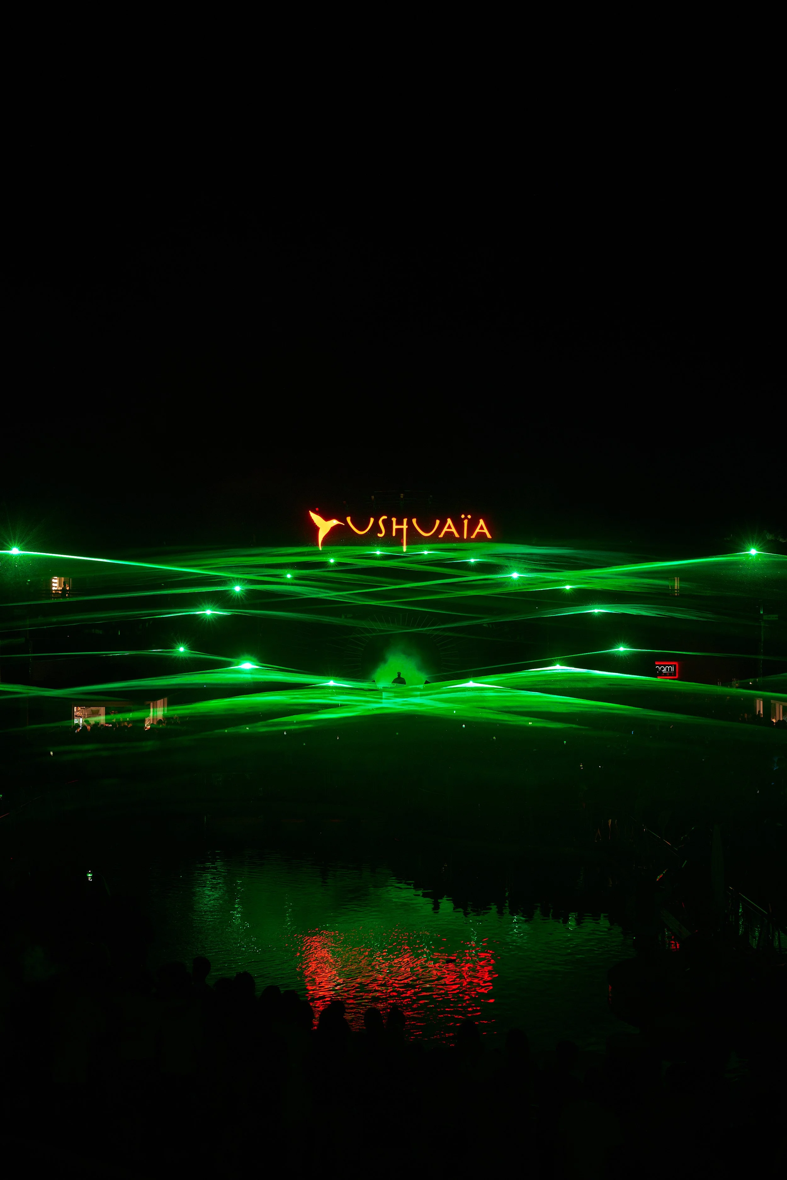 Nighttime water show with green laser lights across a body of water, and a illuminated sign that reads 'Ushuaïa' in red with the Ushuaïa logo, reflected in the water.