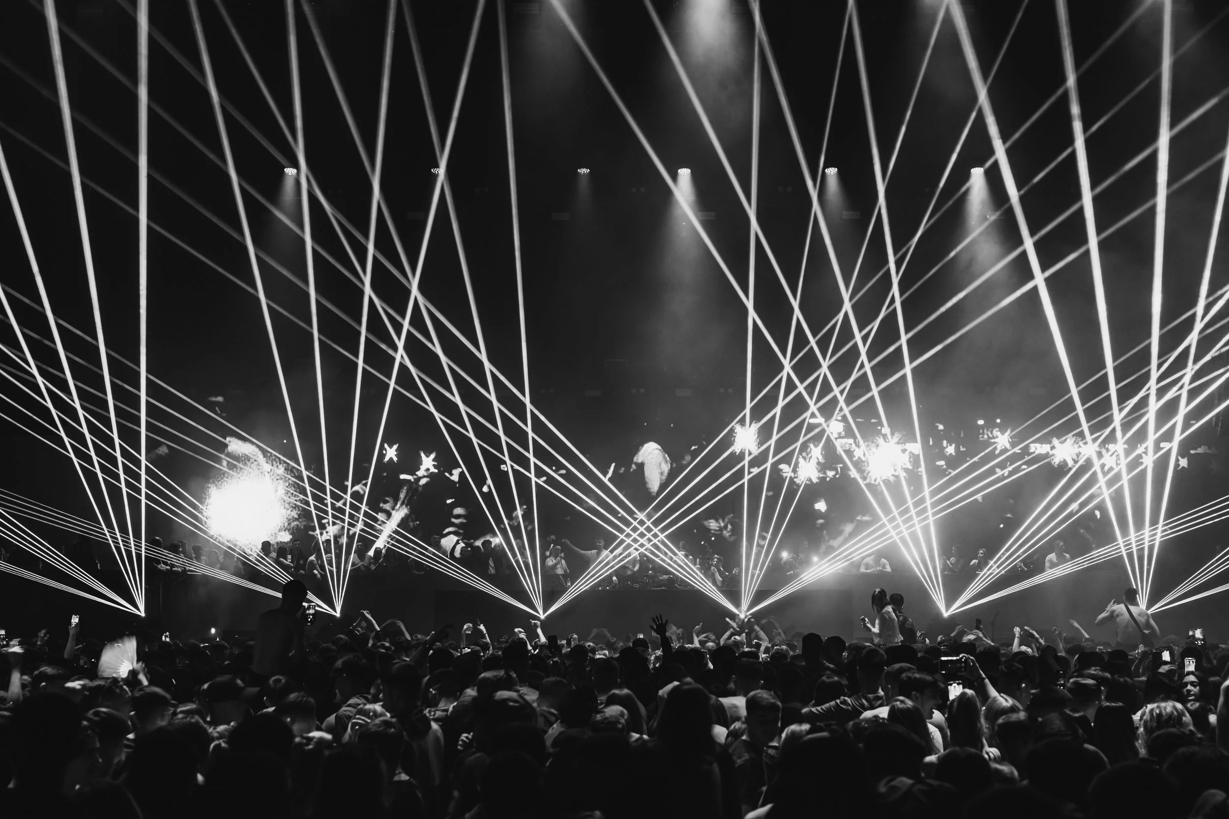 A large crowd of people at a concert or event with bright laser lights and smoke effects in the background.