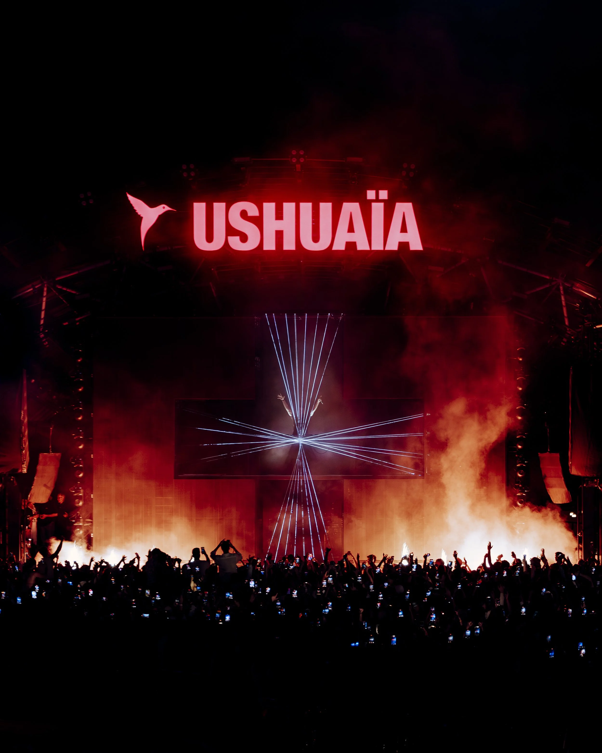 A large crowd attending a concert or festival at night, with a bright red sign displaying the word 'USUAIA' and a hummingbird logo above it, surrounded by colorful lights and smoke.