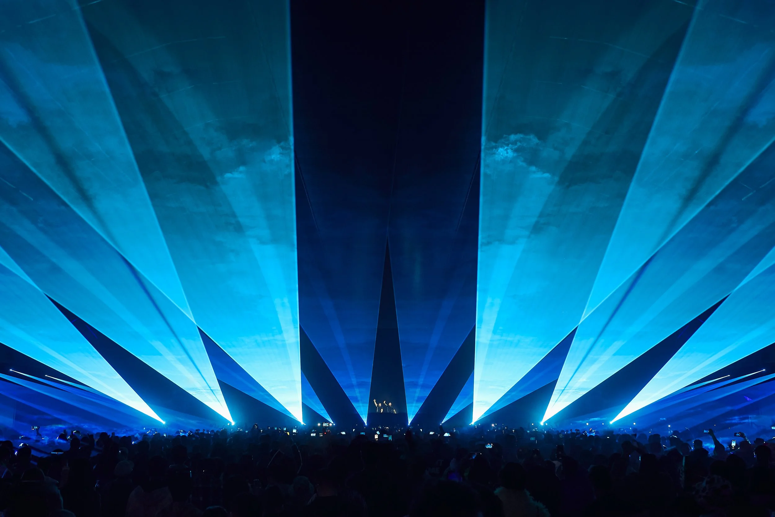 A concert or music event with a large crowd, featuring blue laser lights and a stage with performers.