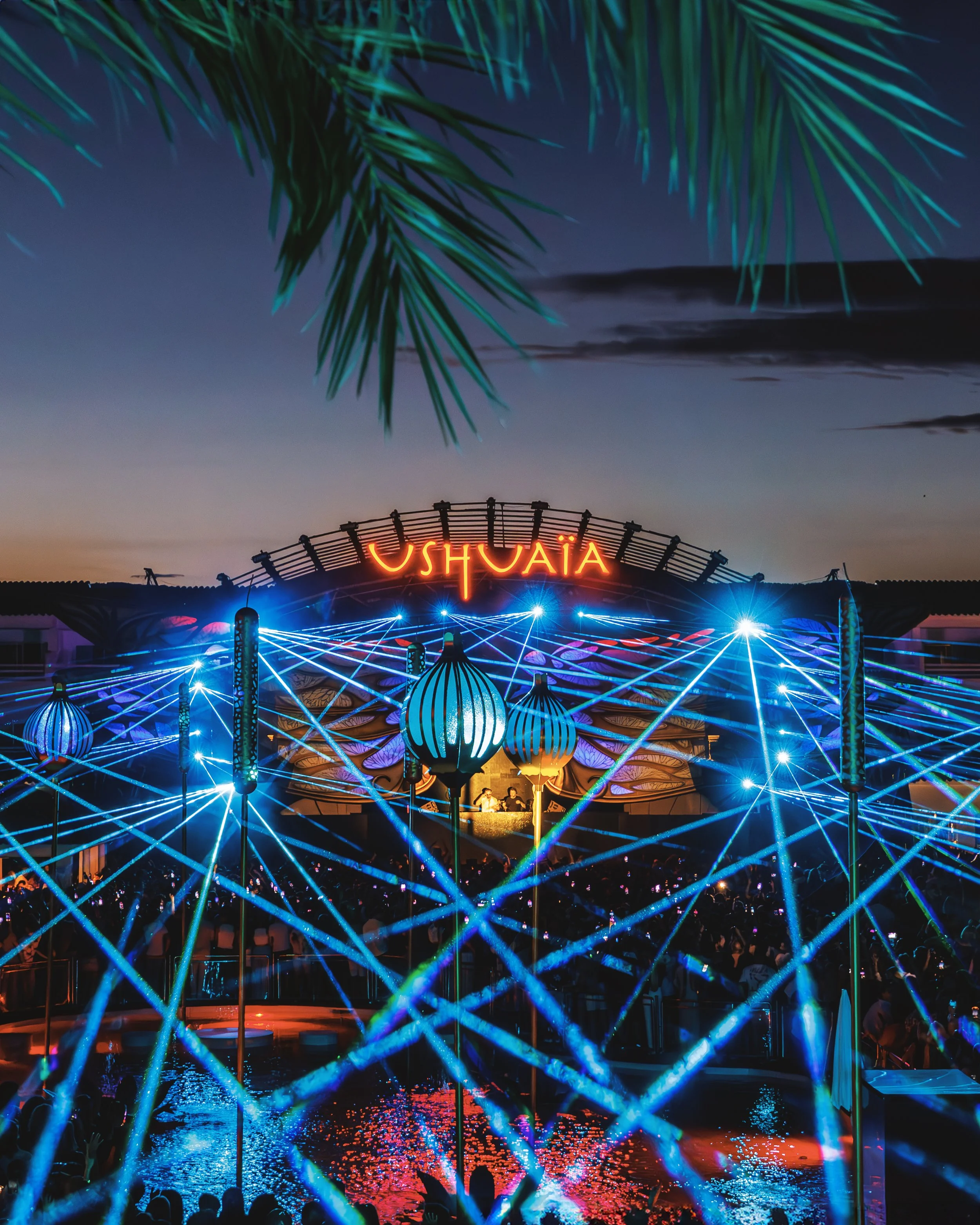 Lighted amusement park ride at night with neon lights, a neon sign reading "Ushuaïa," and palm tree leaves overhead.