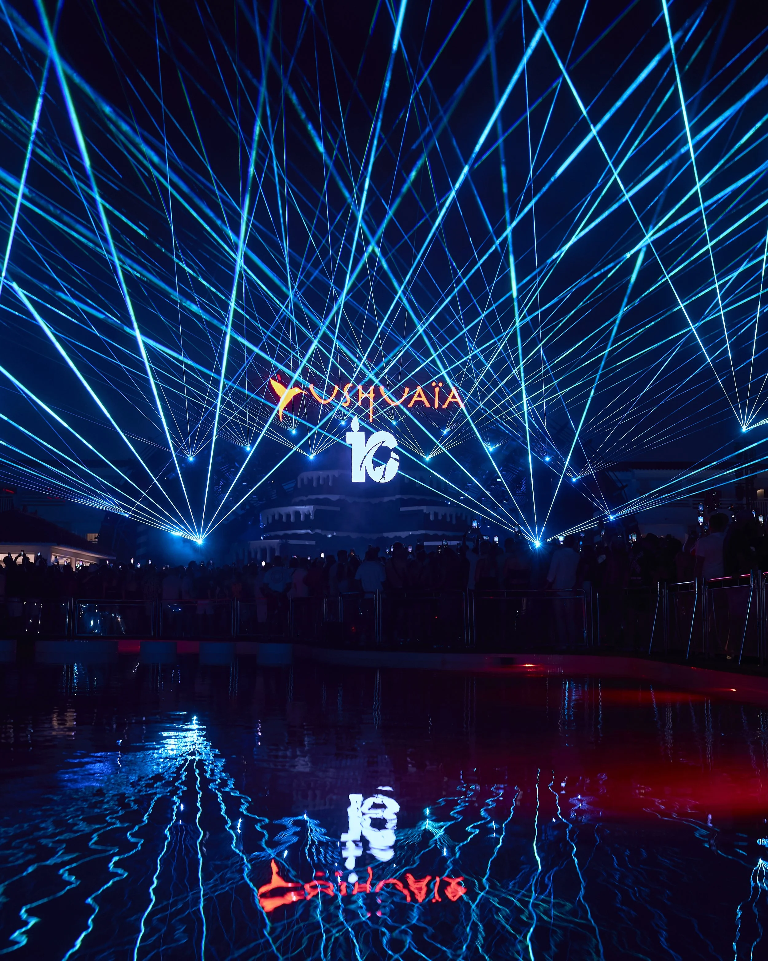 A night scene with a large crowd of people watching a laser light show over a body of water. The laser beams are blue and white, crisscrossing in the sky. A large illuminated sign reads 'FUSHUAIA' in red and orange colors, reflected in the water.