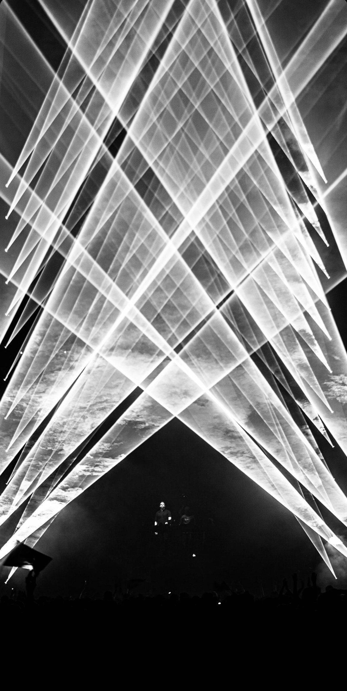 A black and white photo of an indoor laser light show with beams of light forming geometric patterns in the air. There are two people standing on a platform at the bottom of the image, looking up at the display.