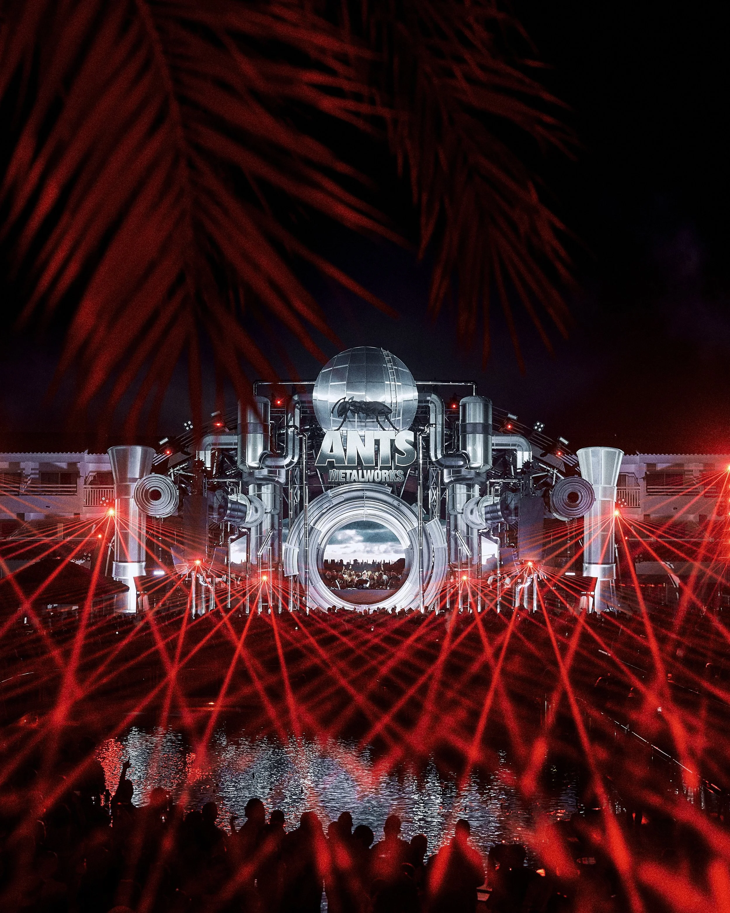 Nighttime festival scene with a crowd of people watching a performance on a stage resembling a giant industrial structure with a large logo that says "ANTS METALWORKS." Fireworks light up the sky above, and the reflection can be seen on the water in 
