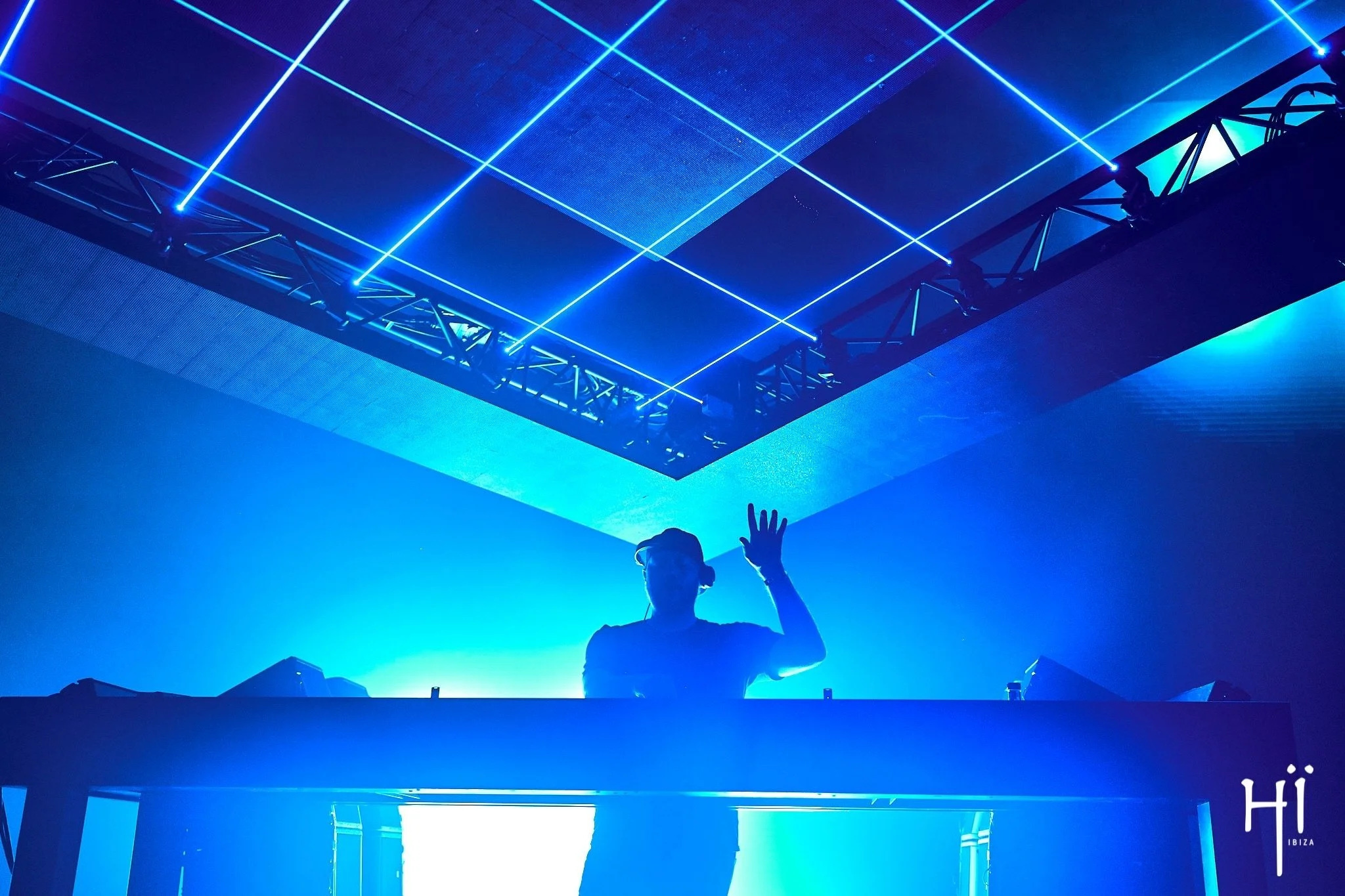 A person standing under a ceiling with blue neon lights arranged in a grid pattern, with their hand raised and the background illuminated in blue tones.