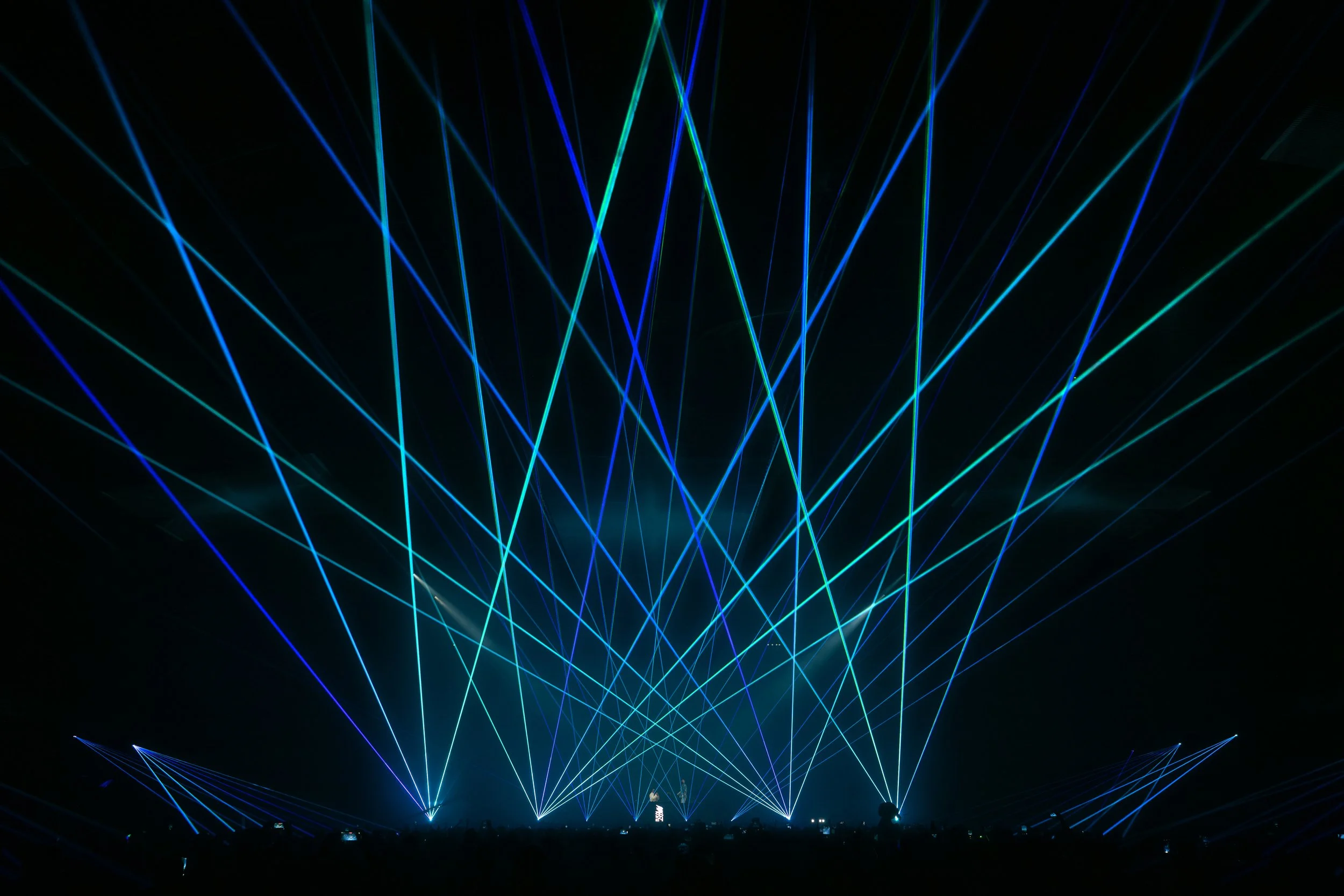 Colorful laser lights and spotlights illuminating a dark concert or event space with a crowd silhouetted in the foreground.