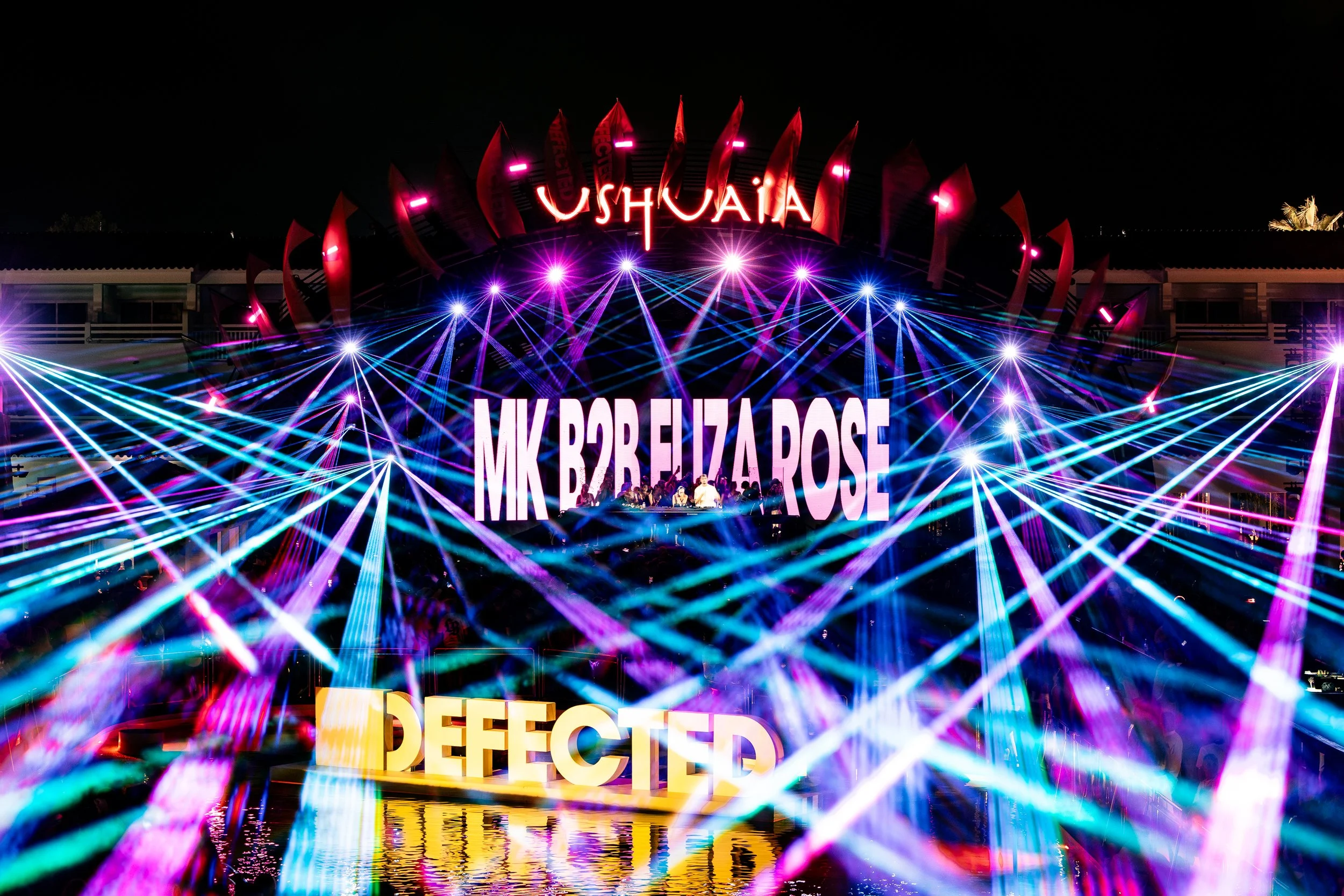 Light display at night with a sign that reads 'Ushuaïa', colorful lights, laser beams, and a stage with people.