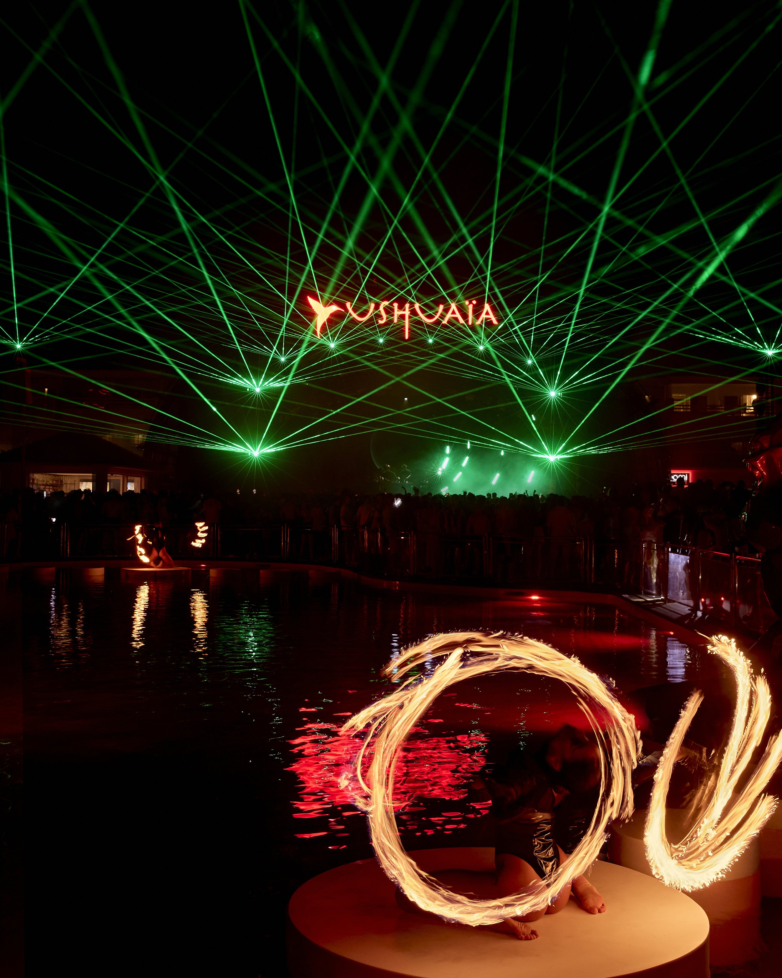 A performer creating fire poi light trails on a circular platform with a backdrop of green laser lights and a neon sign that reads 'YUSHOAIA'.