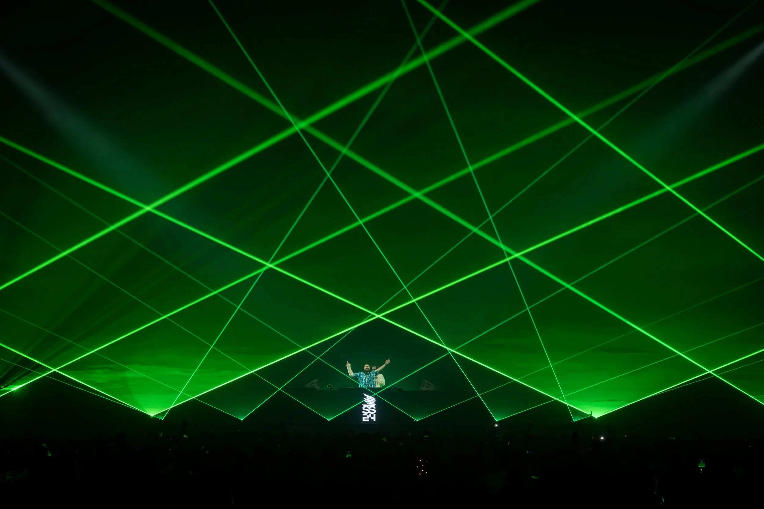 A person on stage with arms raised, surrounded by green laser lights forming intersecting lines in a dark concert or festival setting.