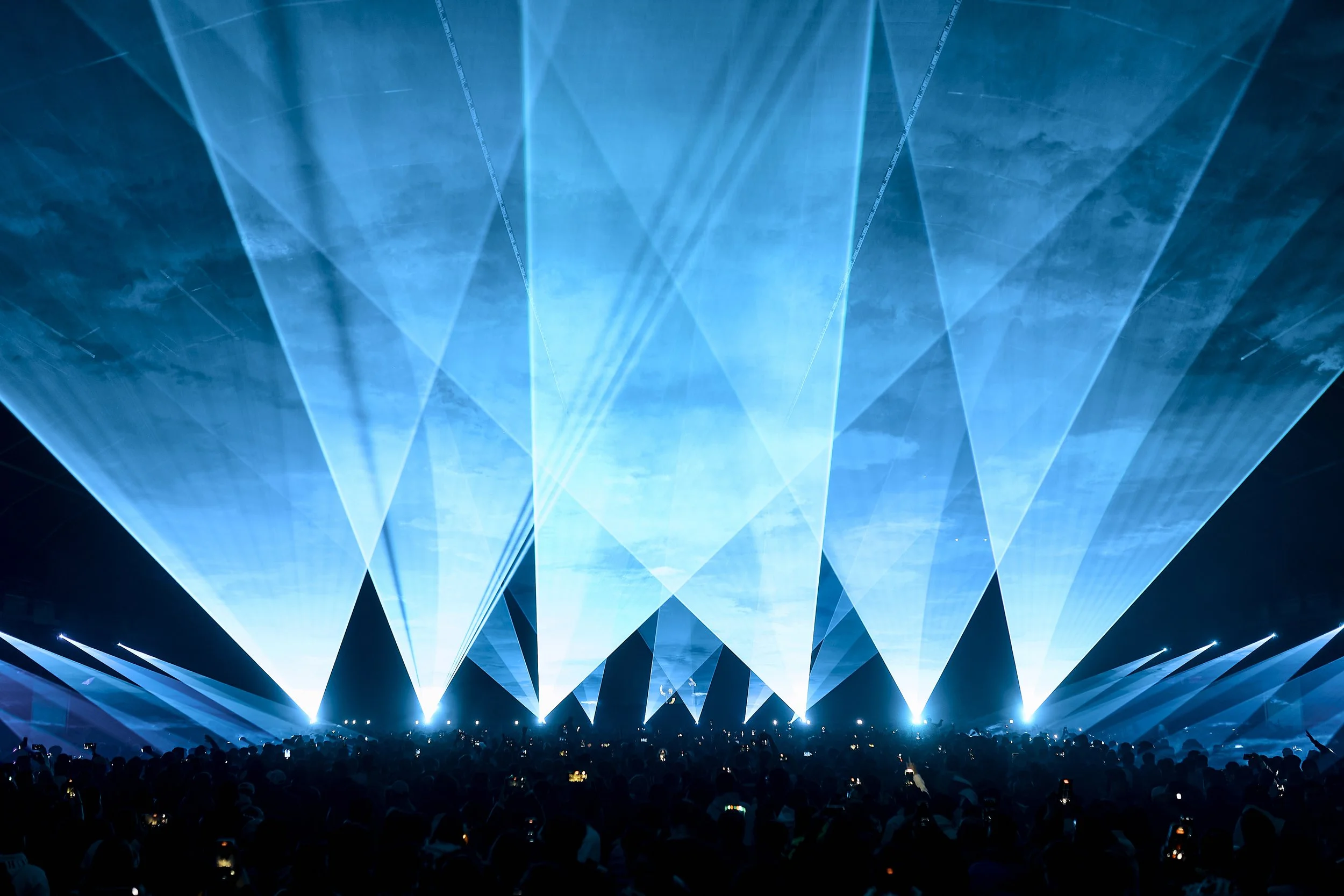 A concert or music event with blue laser lights and beams creating geometric patterns in the air, with a large audience watching from below.