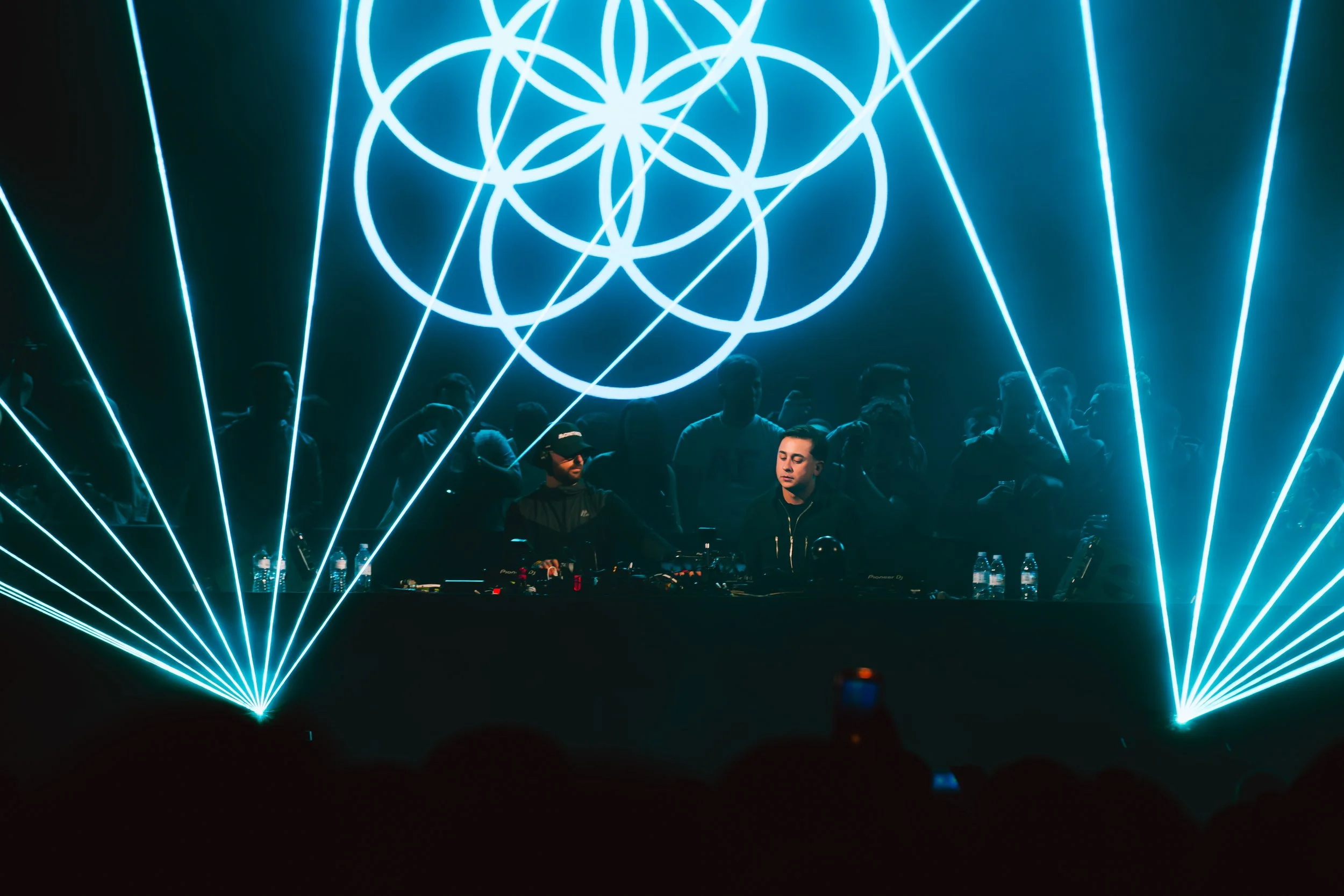 A DJ performs at a dark concert or nightclub with bright blue laser lights creating geometric patterns above and behind him. The DJ is focused on his equipment in front of a crowd of people.
