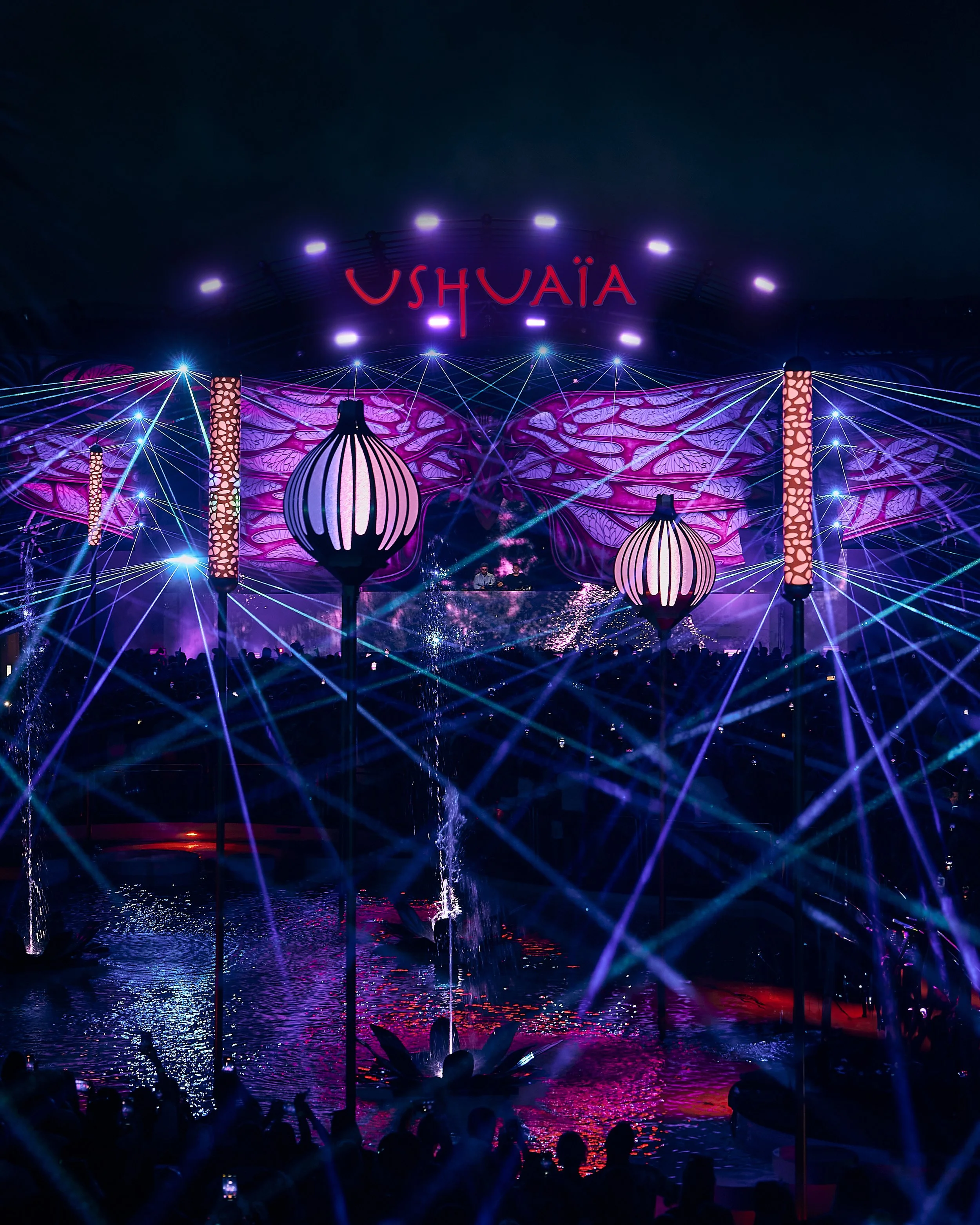 Colorful night scene of a stage or event area with purple, pink, and blue lighting, large hanging lanterns, and a crowd of people in the foreground. The background features a sign that says 'USHUAIA' in red neon letters.