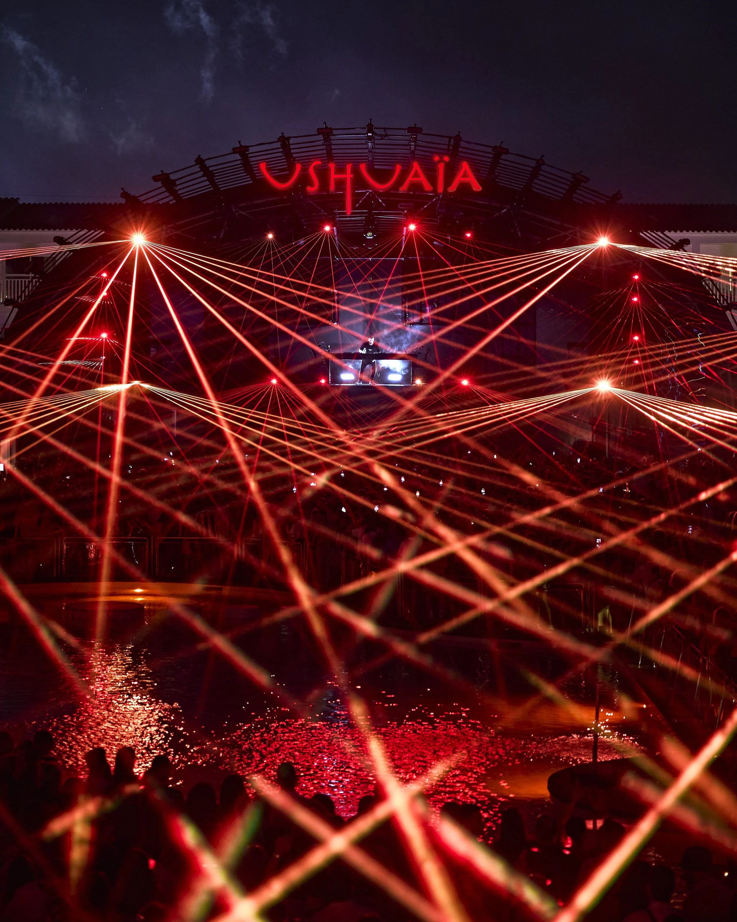 Nighttime scene of a water show with red laser lights, reflections on the water, and silhouettes of people sitting on the shore in Ushuaia, Argentina.