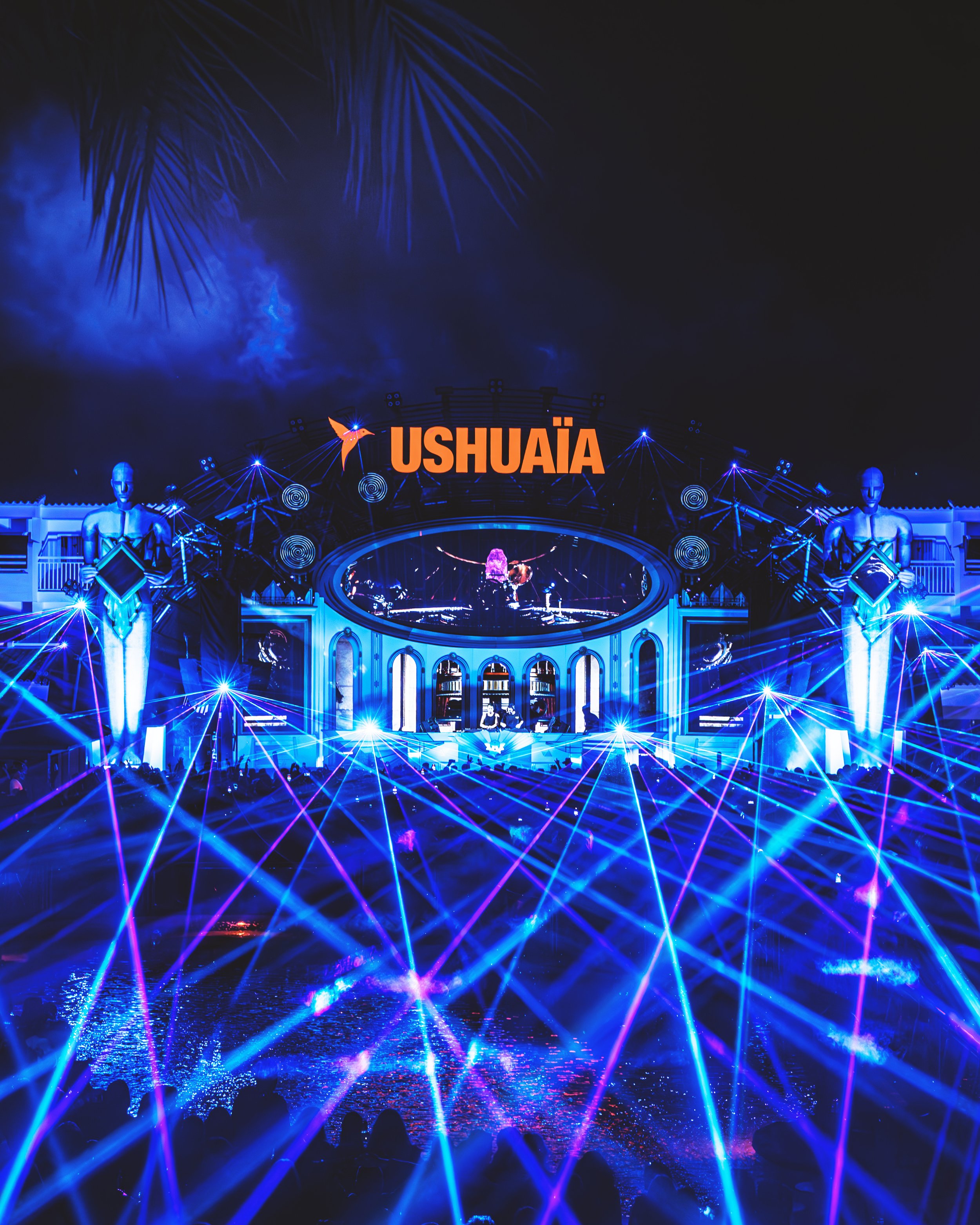 Nighttime scene at Ushuaïa with bright blue laser lights, fireworks in the sky, and silhouettes of people in the foreground.