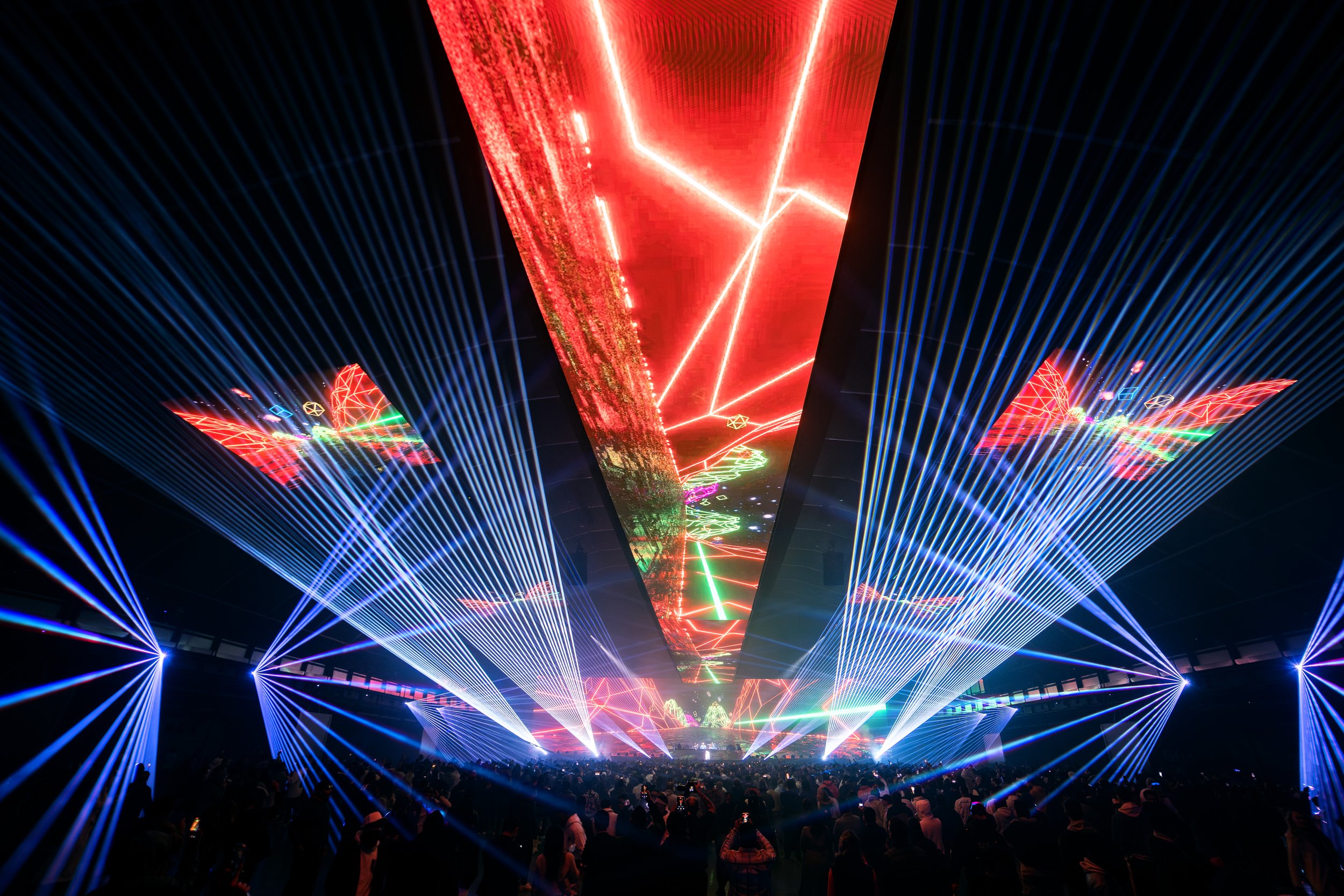 A large crowd attends a concert or music event with colorful laser lights and digital projections on the ceiling.