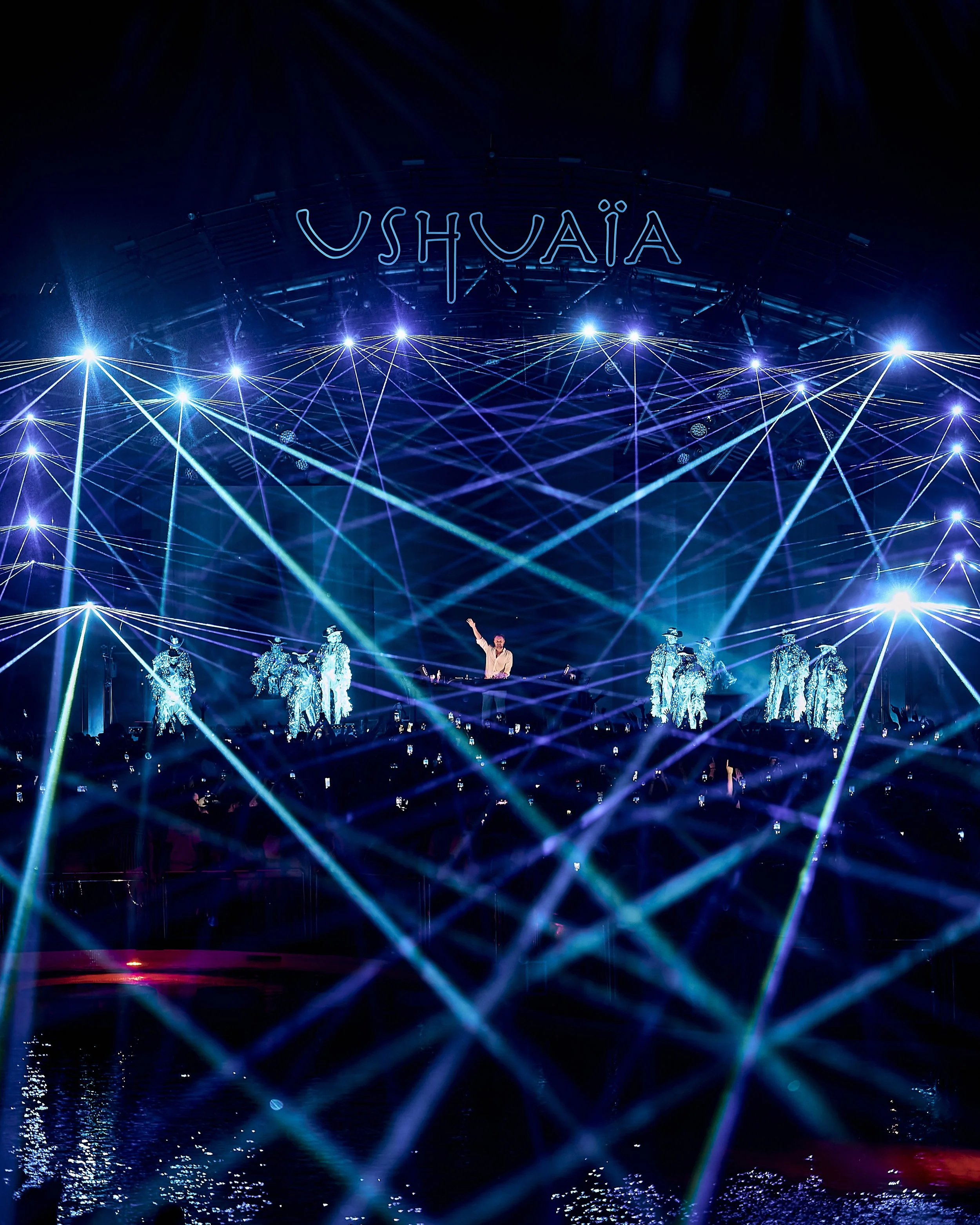 Performance on stage with DJ and dancers in illuminated costumes, surrounded by blue and purple laser lights and a neon sign that reads 'USHUAÏA'