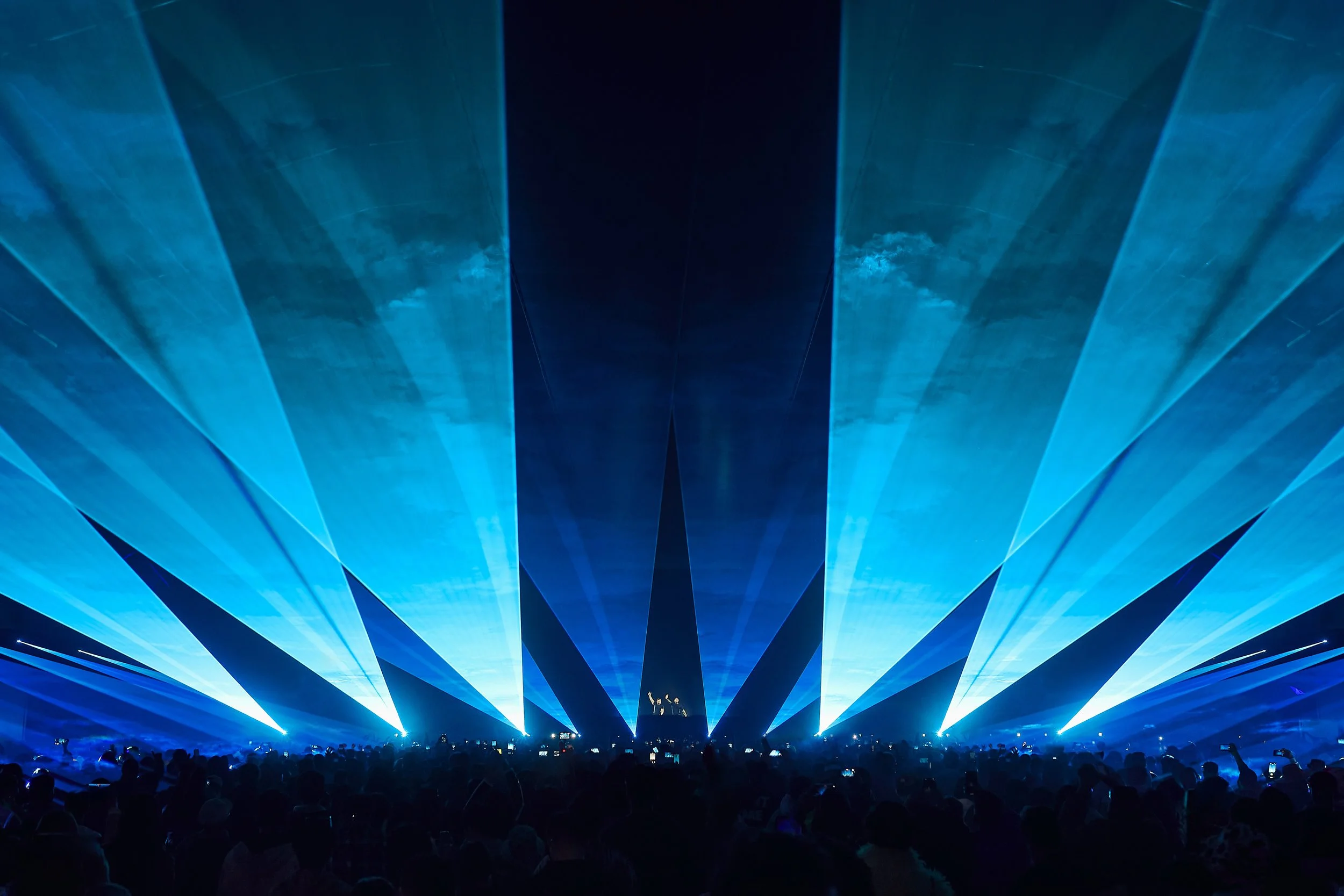 Large crowd at a concert with blue laser lights projecting beams into the sky, creating a dramatic visual effect.