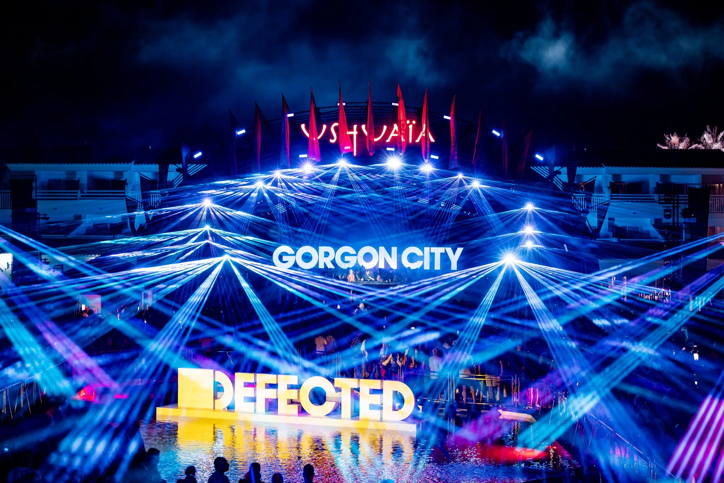 Nighttime scene at Gorgon City concert with vibrant blue and purple laser lights, a large illuminated sign reading 'GORGON CITY', and the word 'PERFECTED' in bold yellow letters at the front.