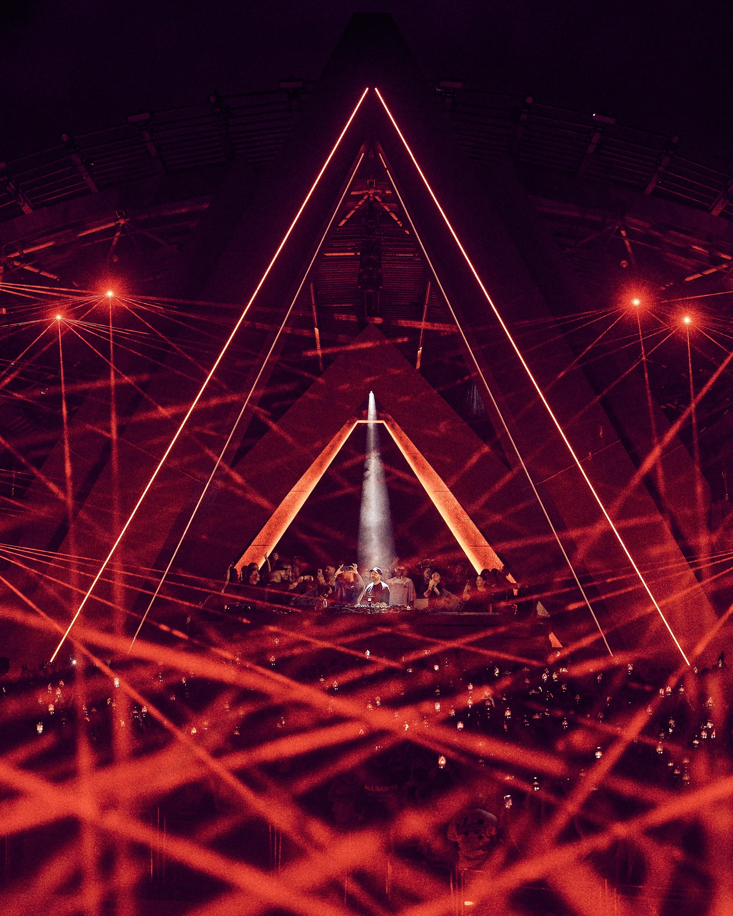 Nighttime scene of a large concert stage with red laser lights forming geometric patterns, and a DJ performing in the center with a crowd watching.