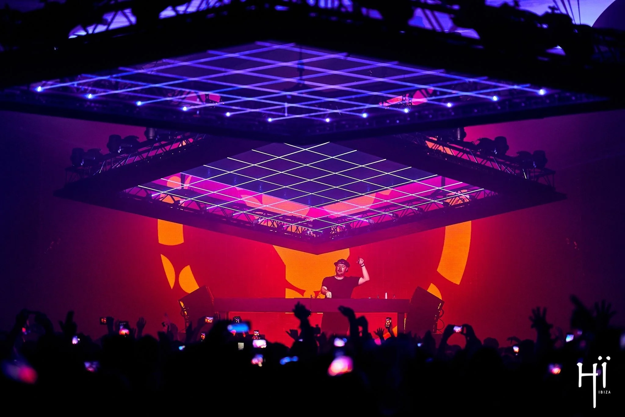A DJ performs on stage under colorful LED light structures during a concert at Hï Ibiza. Crowd members hold up phones and hands in the foreground.