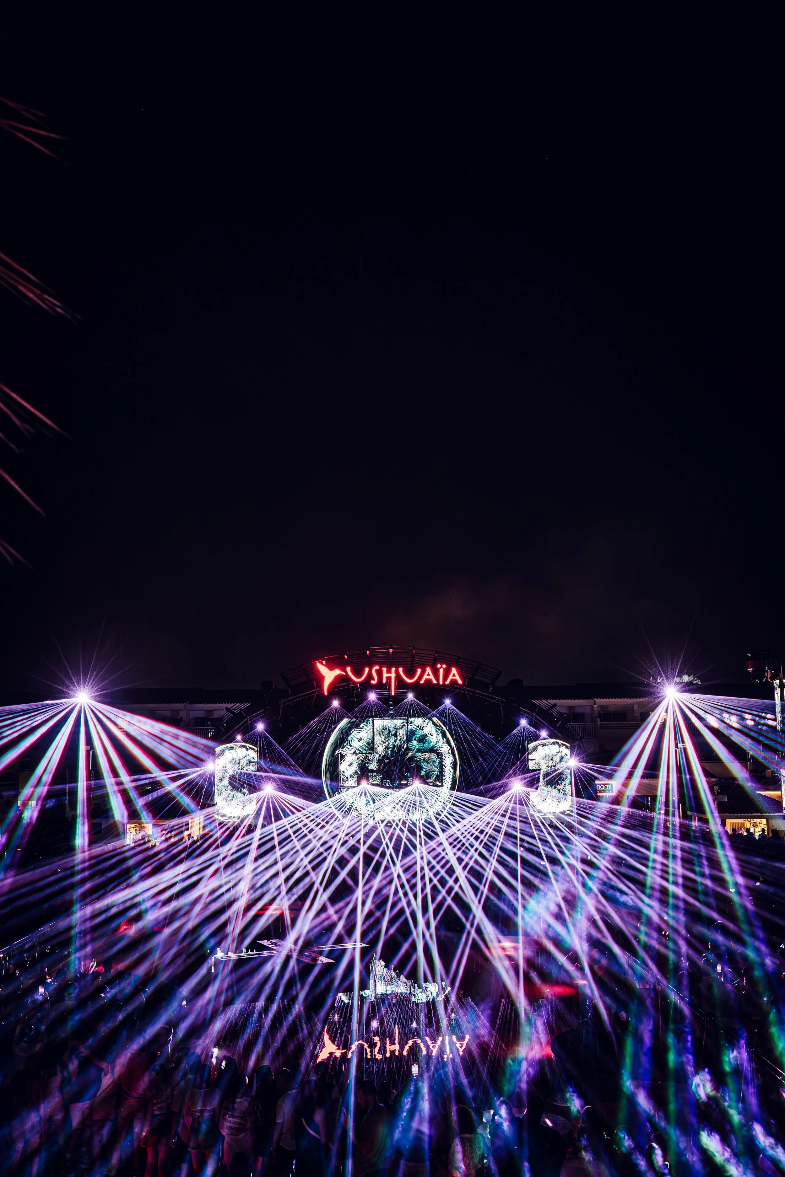 Nighttime scene at the Ushuaia music venue featuring colorful laser lights, crowd silhouettes, and a large glowing display with the venue's name.