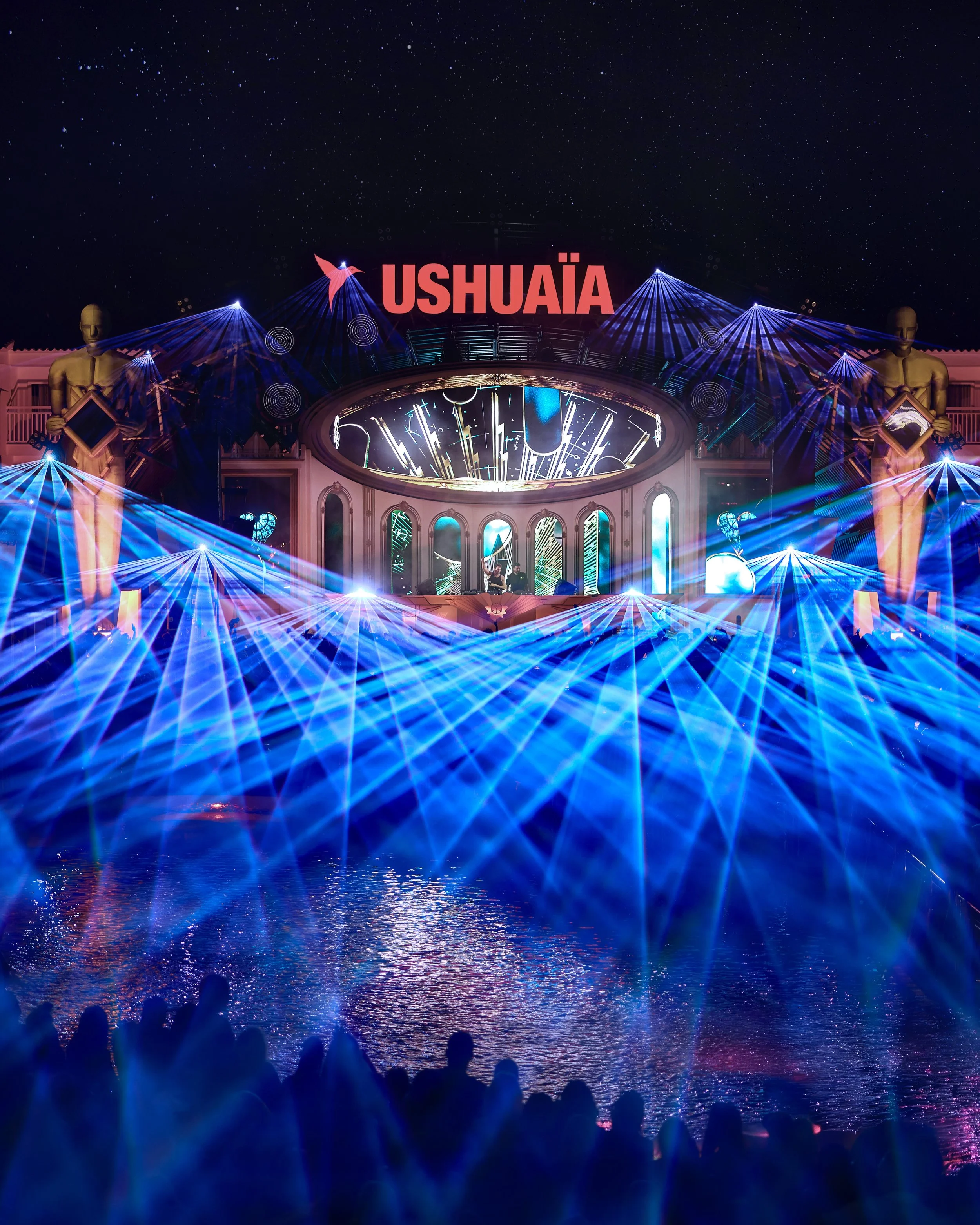 Nighttime scene of a stage with bright lights, statues, and the large red sign reading 'Ushuaïa'. The stage is reflected in water with silhouettes of people watching.