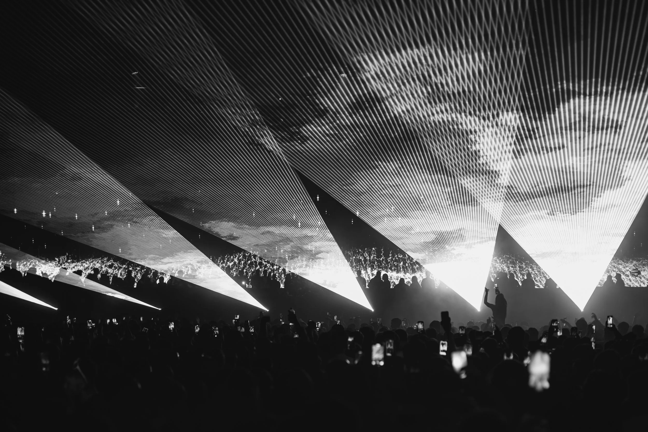 Black and white photo of a large crowd at a concert or event, with silhouettes of people in the foreground and a stage with bright lights and laser effects overhead.