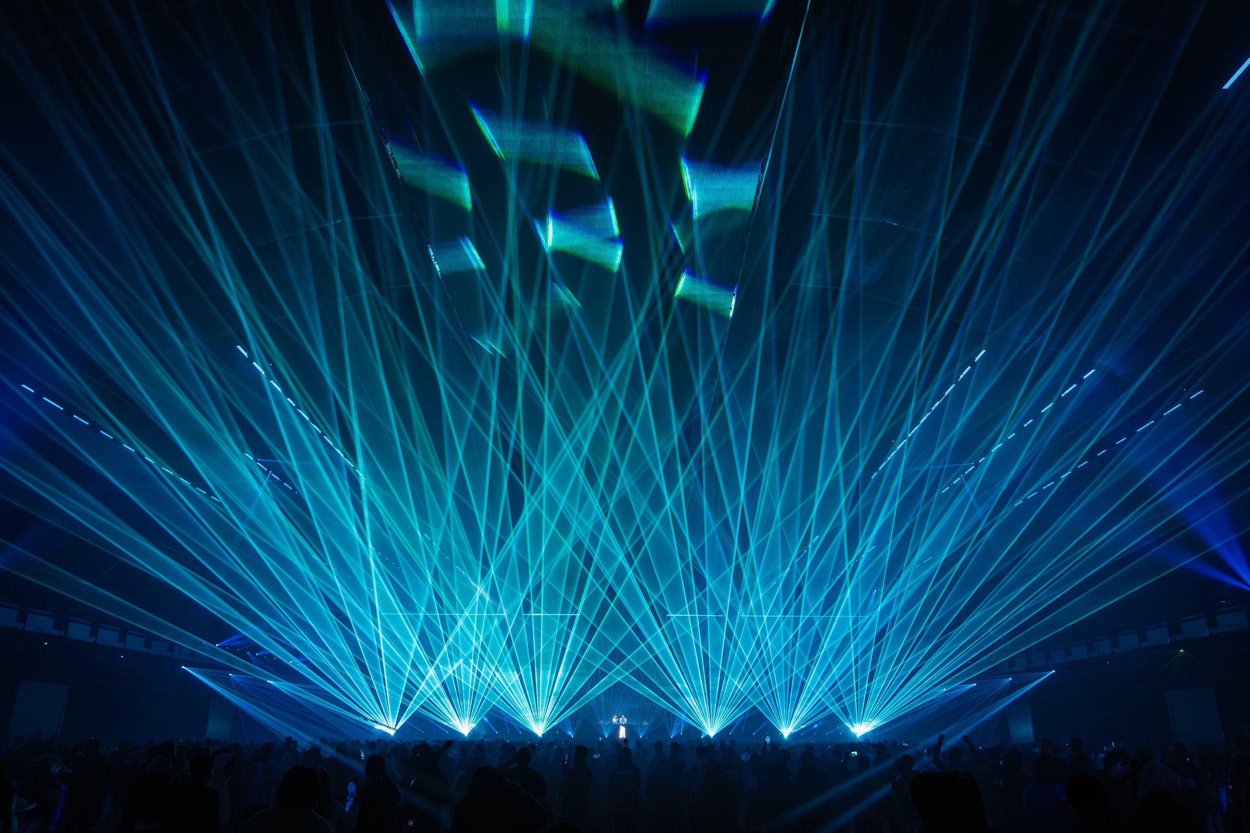 A dark concert venue with blue and green laser lights projected across the room and a crowd of people watching.