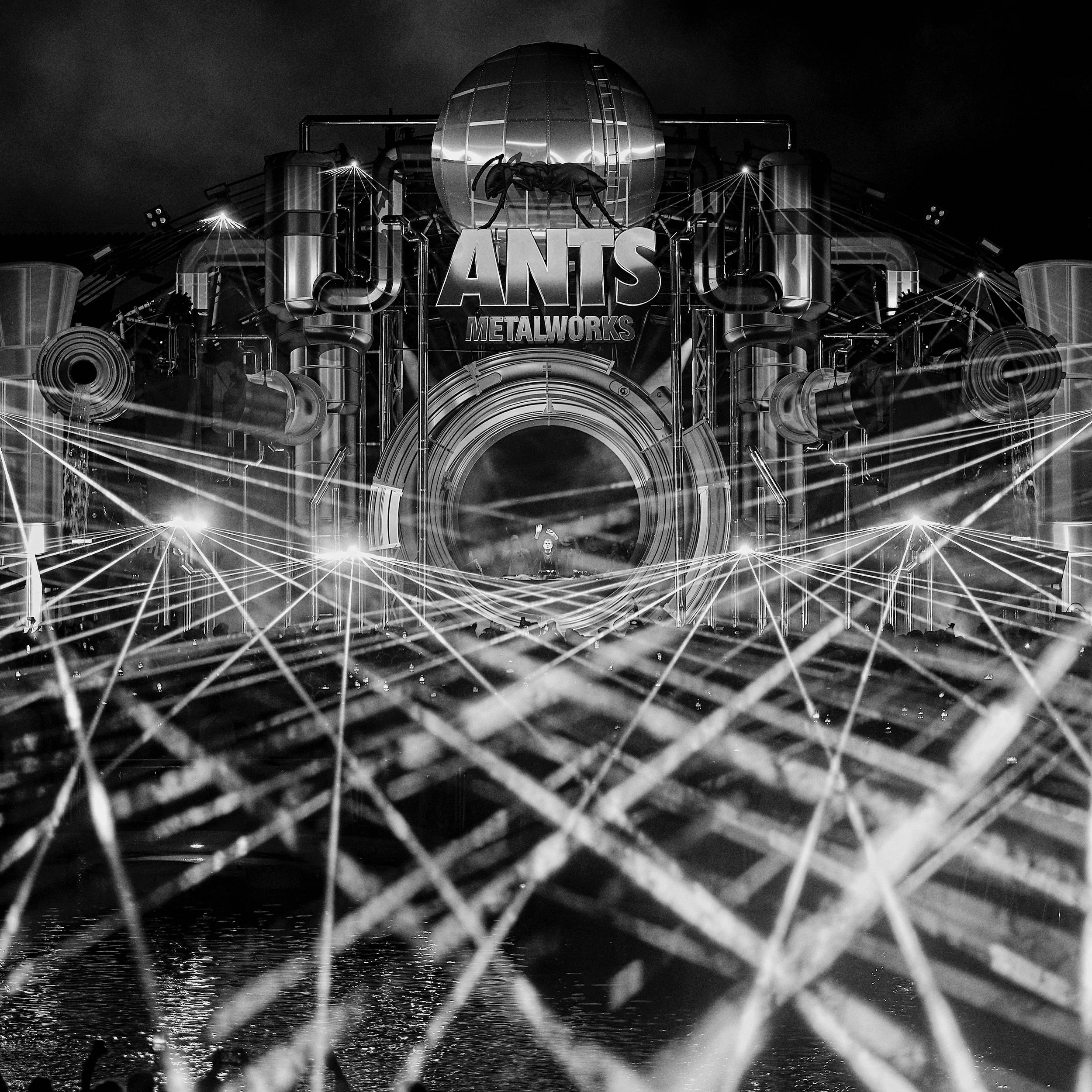 A dark metallic structure with pipes and tubes, with a large sign reading 'ANTIS METALWORKS' and a giant ant sculpture above it, illuminated by bright lights at night.