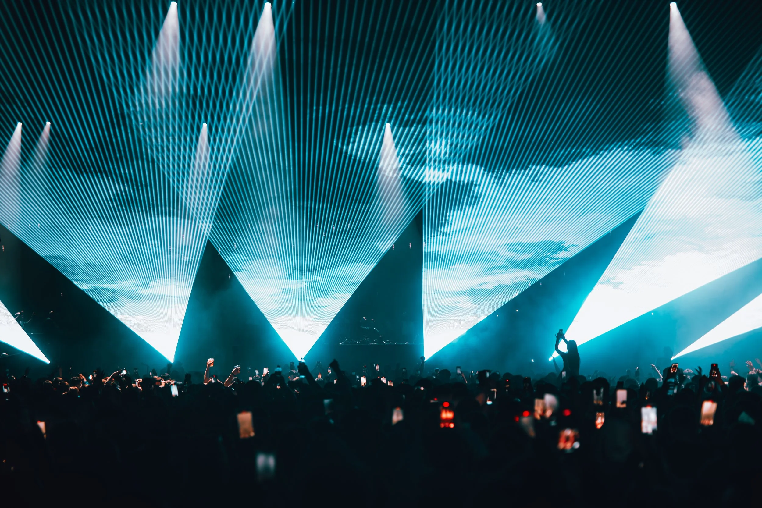 A concert scene with bright blue laser light display overhead and a crowd of people holding up their phones in the dark.