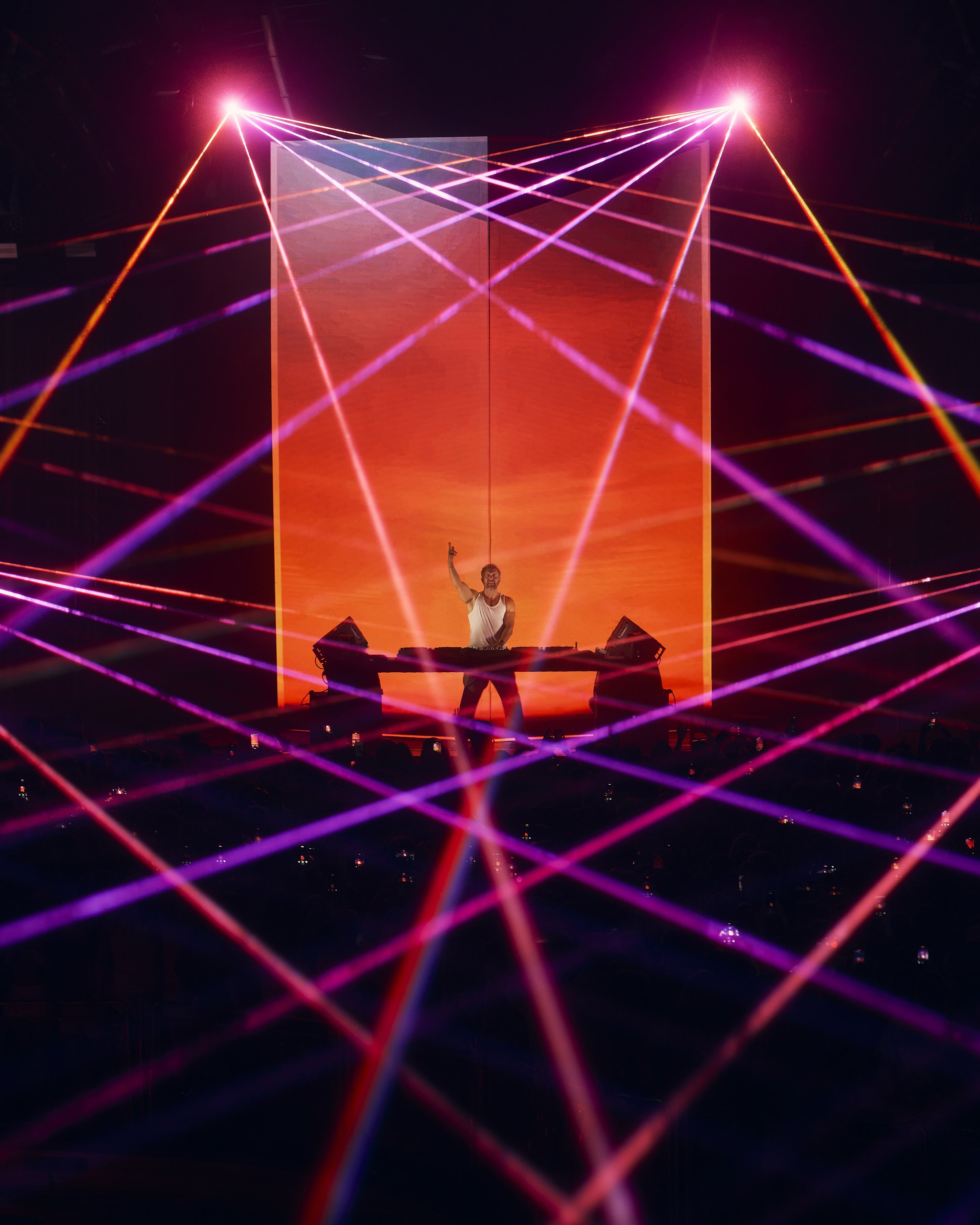 A DJ performing on stage with colorful laser lights forming a geometric pattern, orange and purple hues, and a large orange screen behind him.