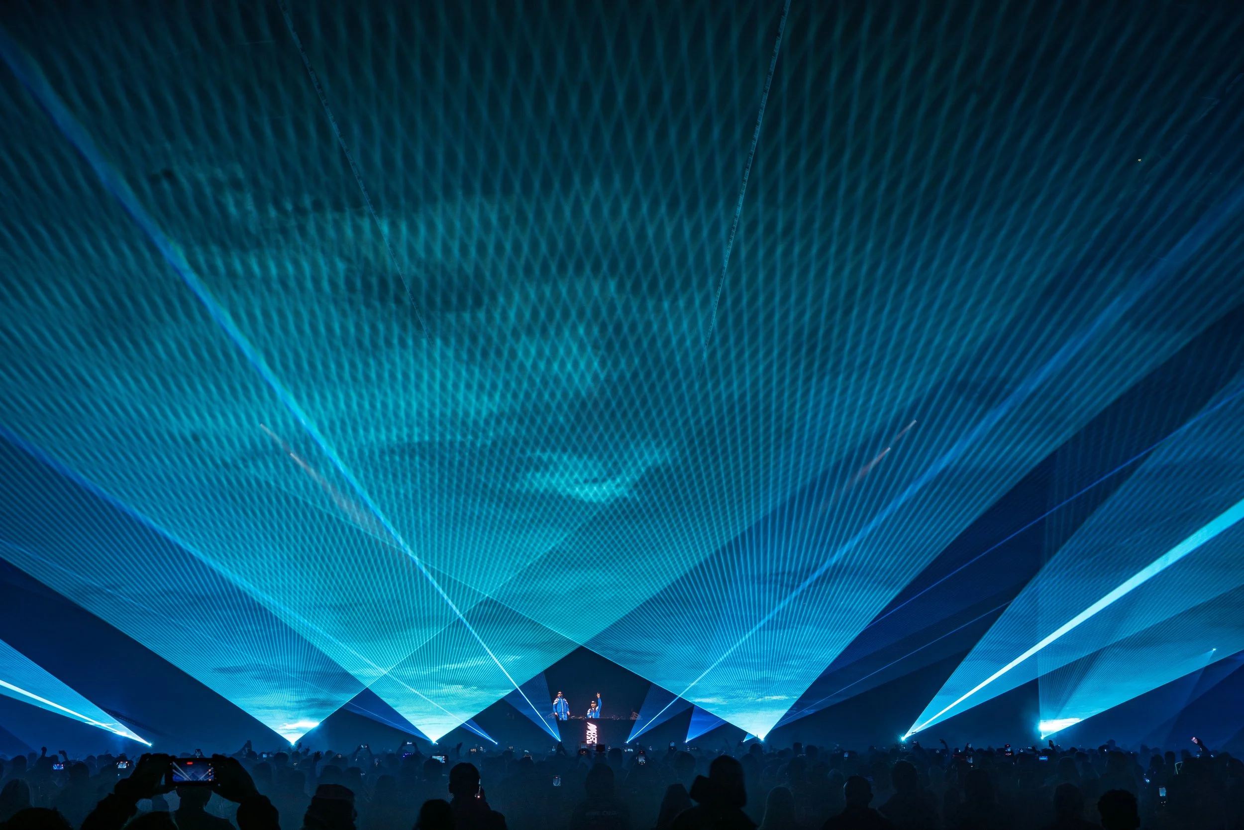 A large indoor concert with a blue laser light show on the ceiling, a stage at the front, and audience members taking photos.