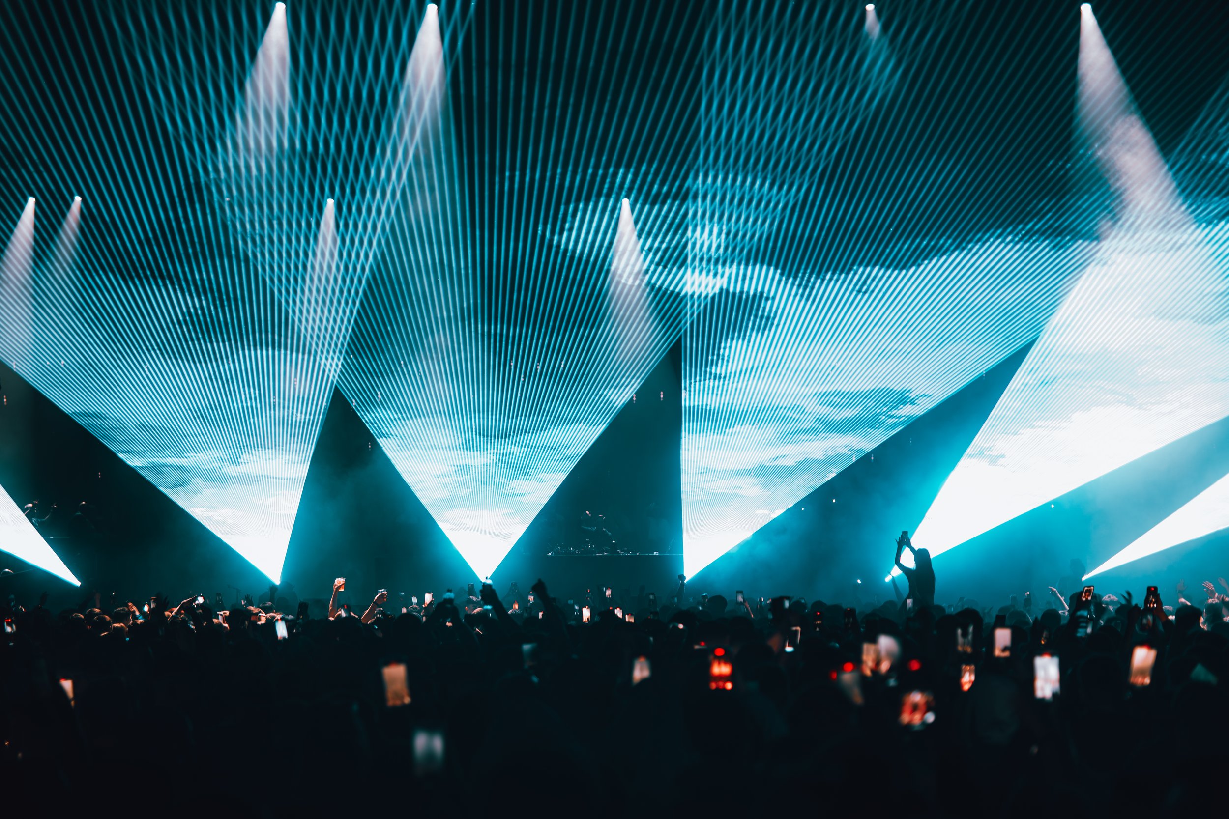 A concert scene with a crowd of people taking photos and videos, illuminated by a large light show with teal and white laser lights forming geometric patterns overhead.