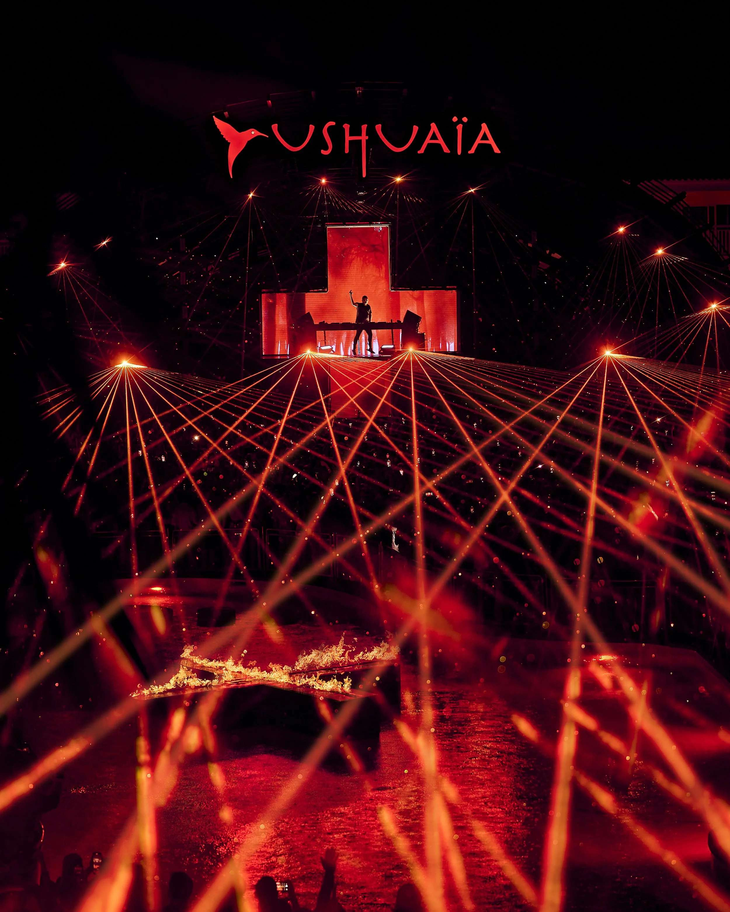 Fireworks display over water with silhouettes of people watching, large illuminated sign spelling 'USHUAÏA' at the top, and a person DJing on stage in front of a red glowing cross-shaped screen.
