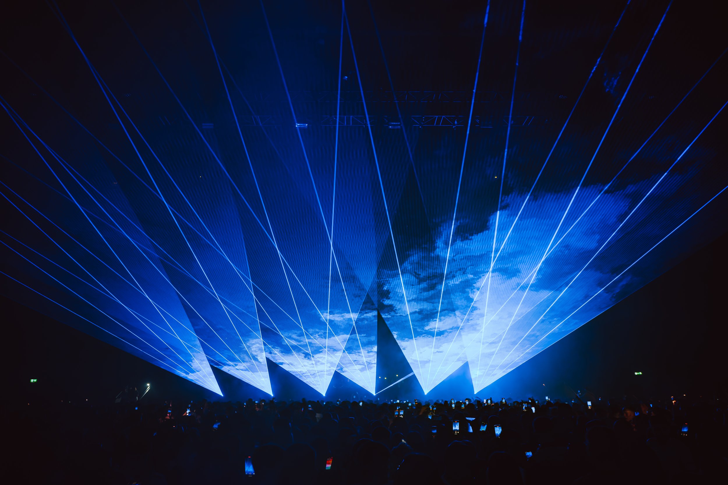 A dark indoor concert with a light display projected on a screen that resembles Earth, surrounded by blue laser lights forming geometric patterns.