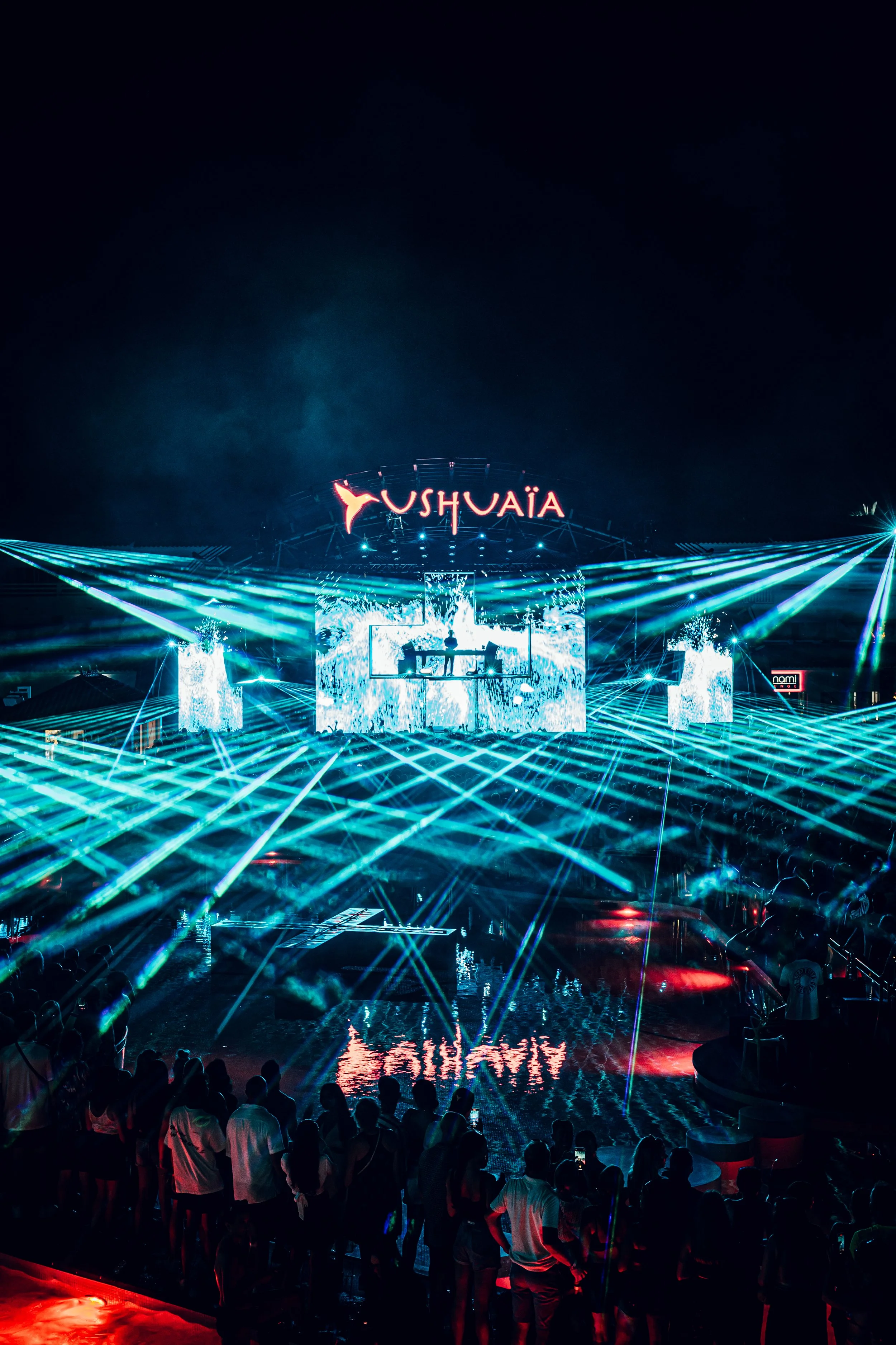Nighttime scene at a music festival or concert with a large crowd, vibrant laser light display, and a stage with a sign reading 'USHUAÏA'.