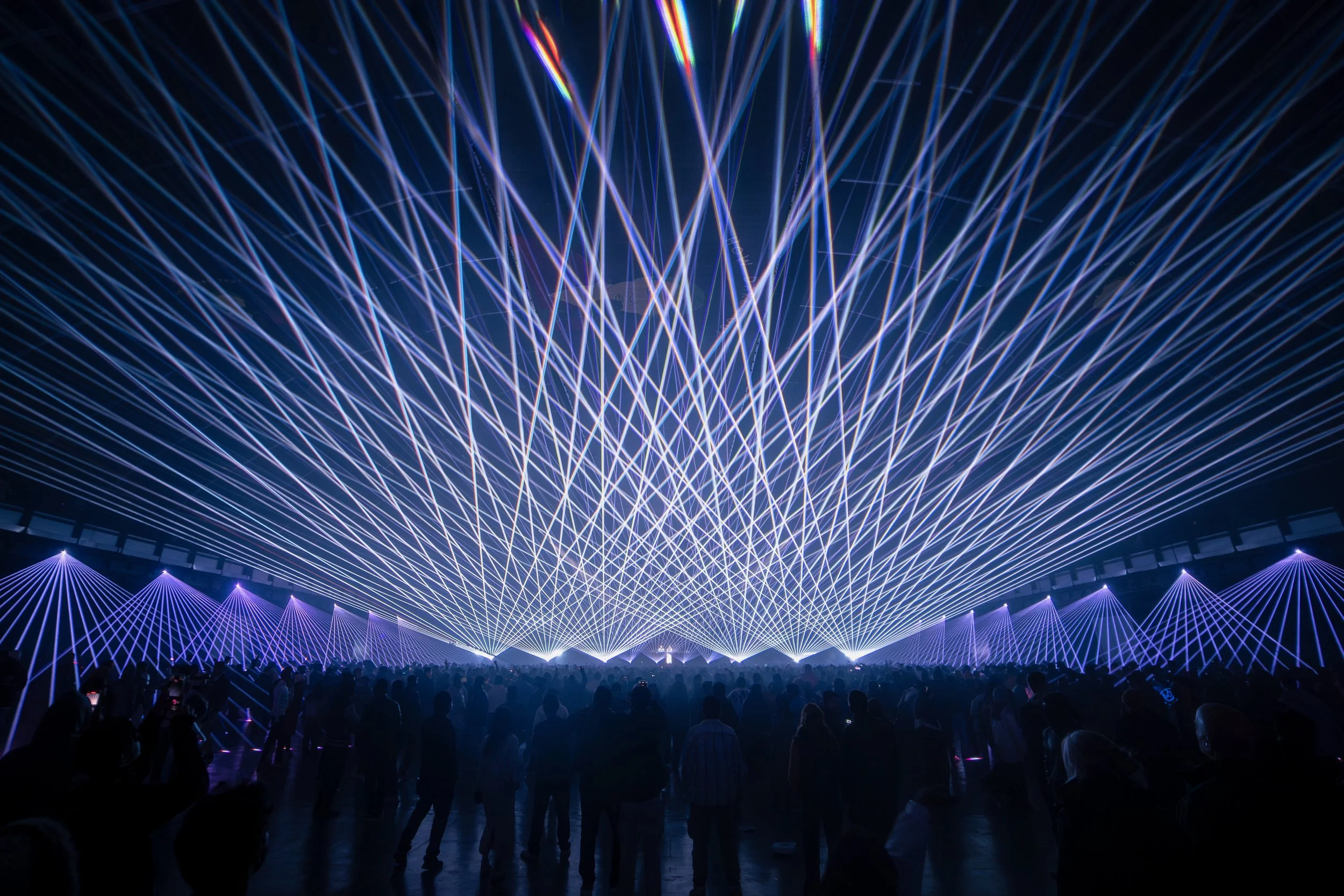 A large crowd watching a laser light show with beams of blue, purple, and red light creating intricate patterns overhead in a dark indoor venue.