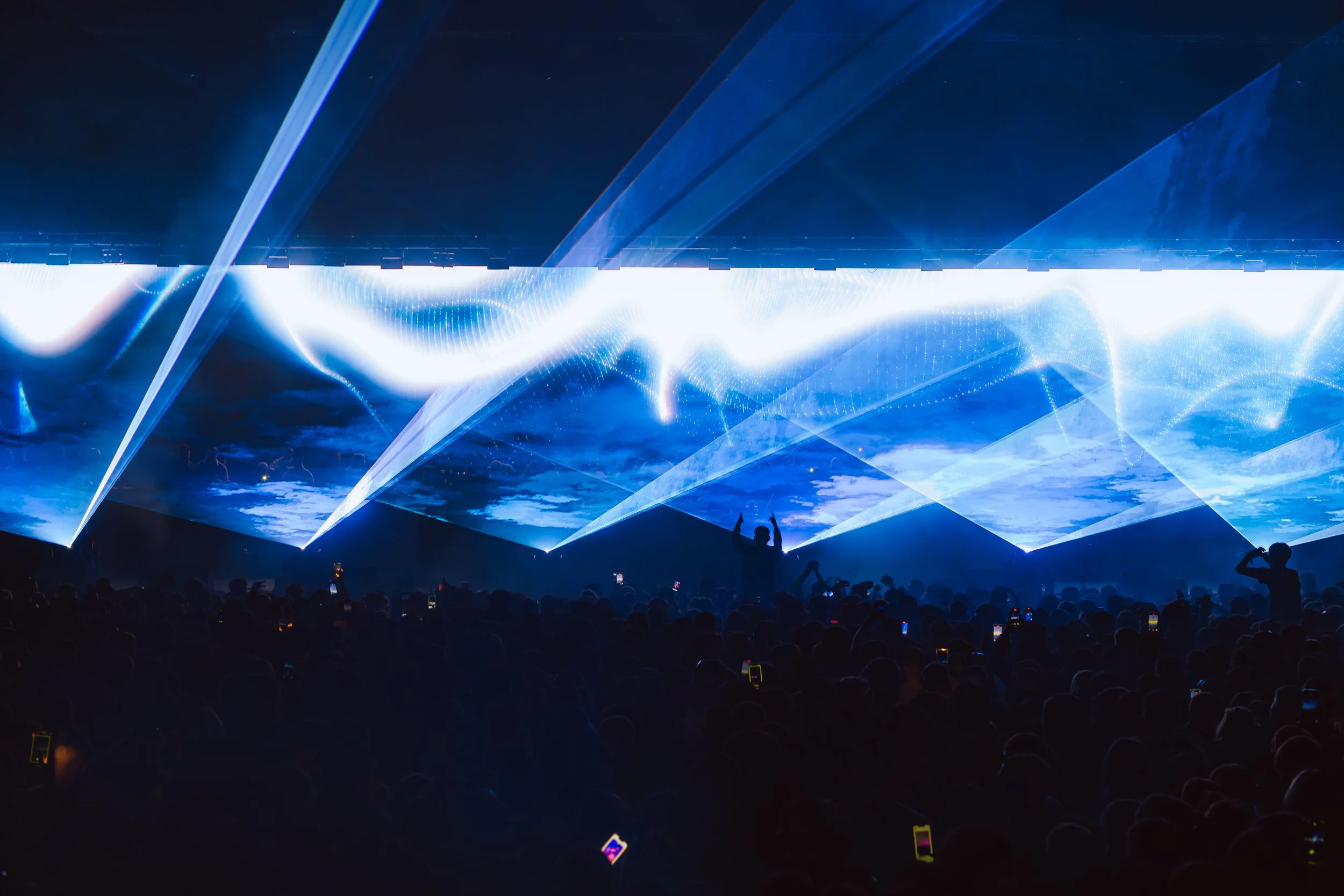 A concert or music festival scene with a large crowd of people watching a light show or laser display with blue and white lights, and some people in the audience are holding up their phones.