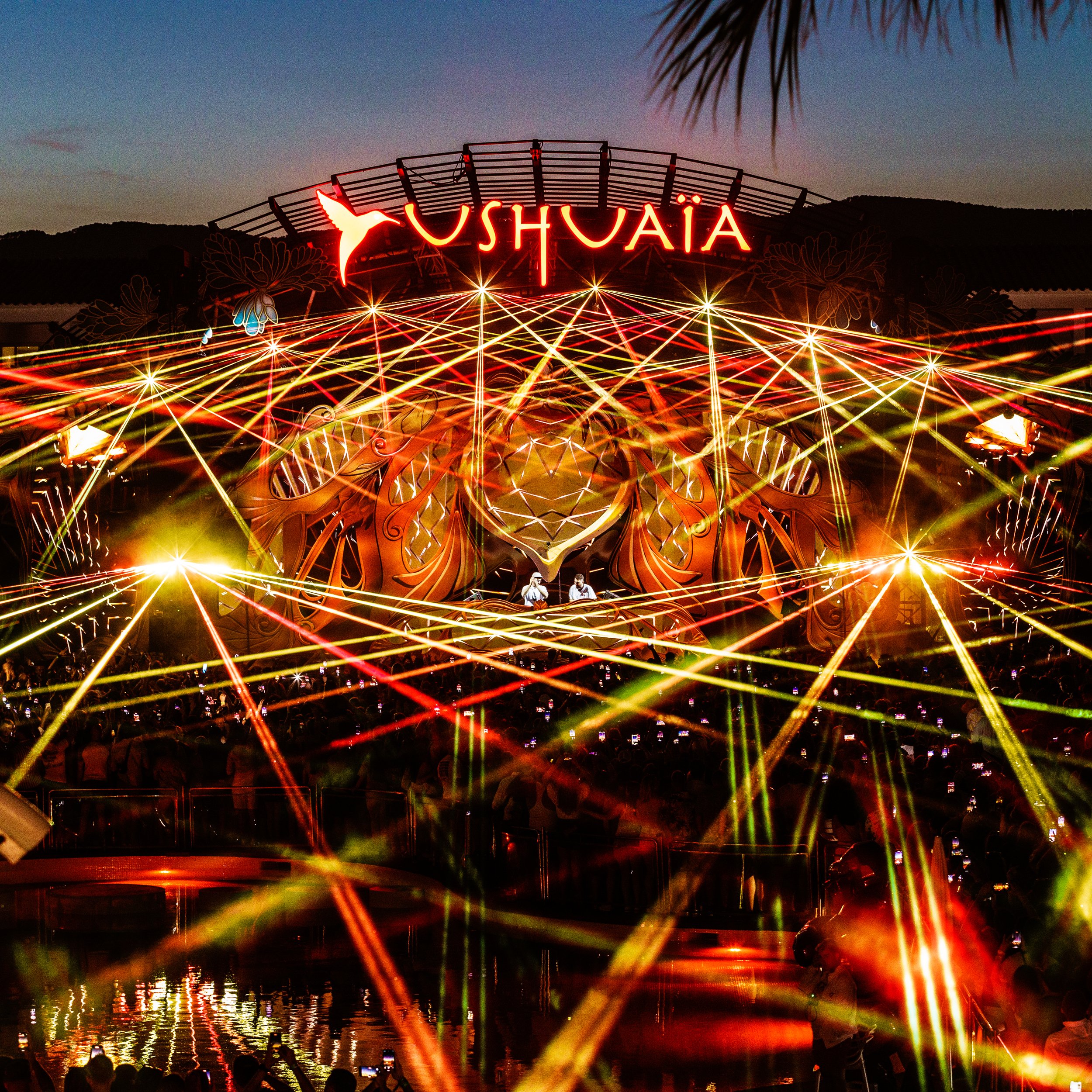 Colorful amusement park ride at sunset with streaks of lights, a large illuminated sign reading 'YUSHUAIA' with a hummingbird logo, surrounded by a crowd of people.