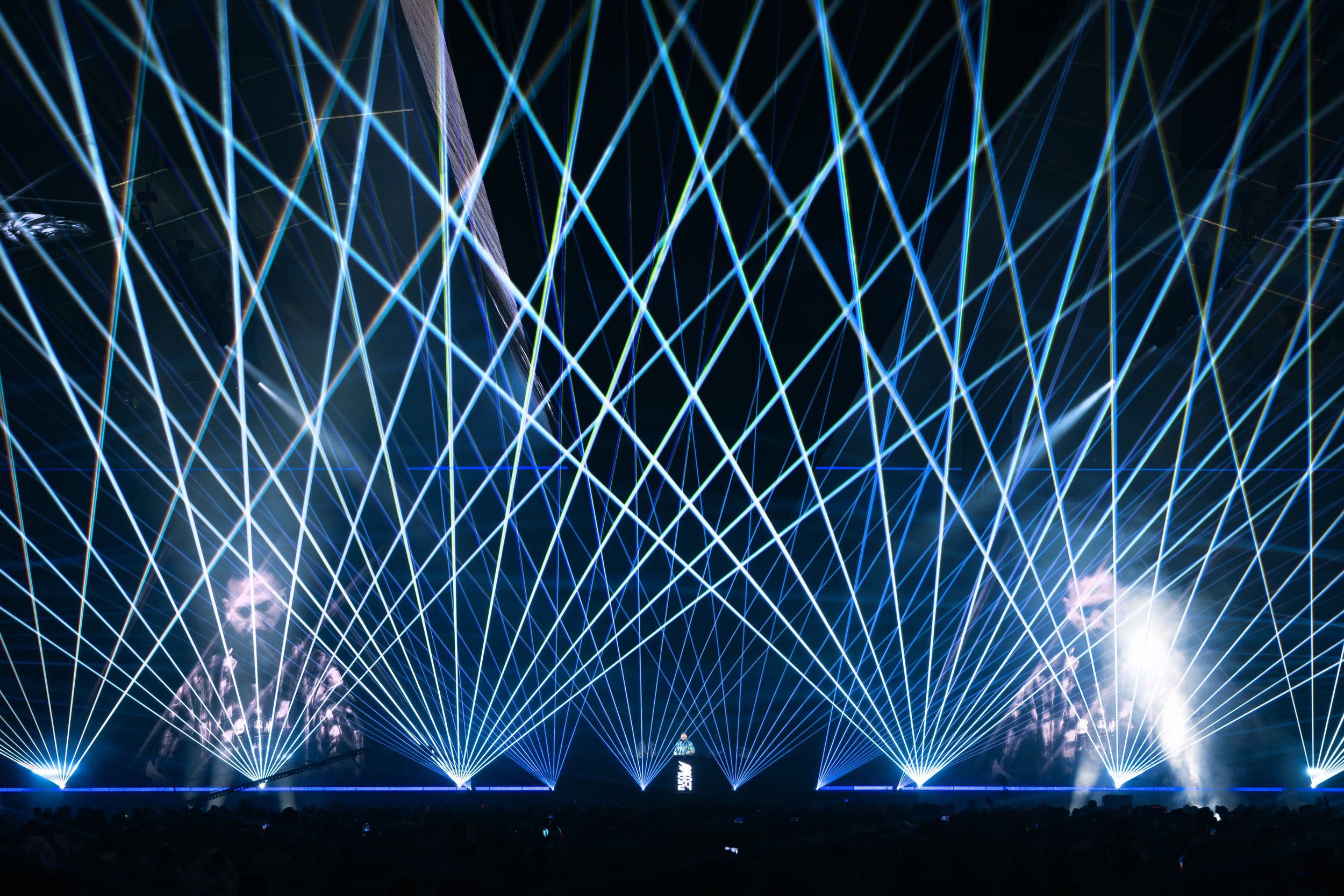 Nighttime laser light show with multiple blue, green, and white laser beams creating intricate patterns above a crowd at a concert or event.