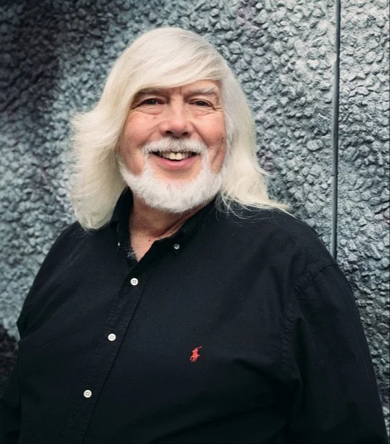 A smiling elderly man with long white hair and a beard, wearing a black shirt with a small red logo, standing against a textured outdoor wall.