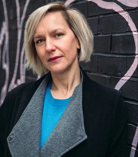 A woman with short blonde hair looking at the camera, standing in front of a black brick wall with pinkish designs.
