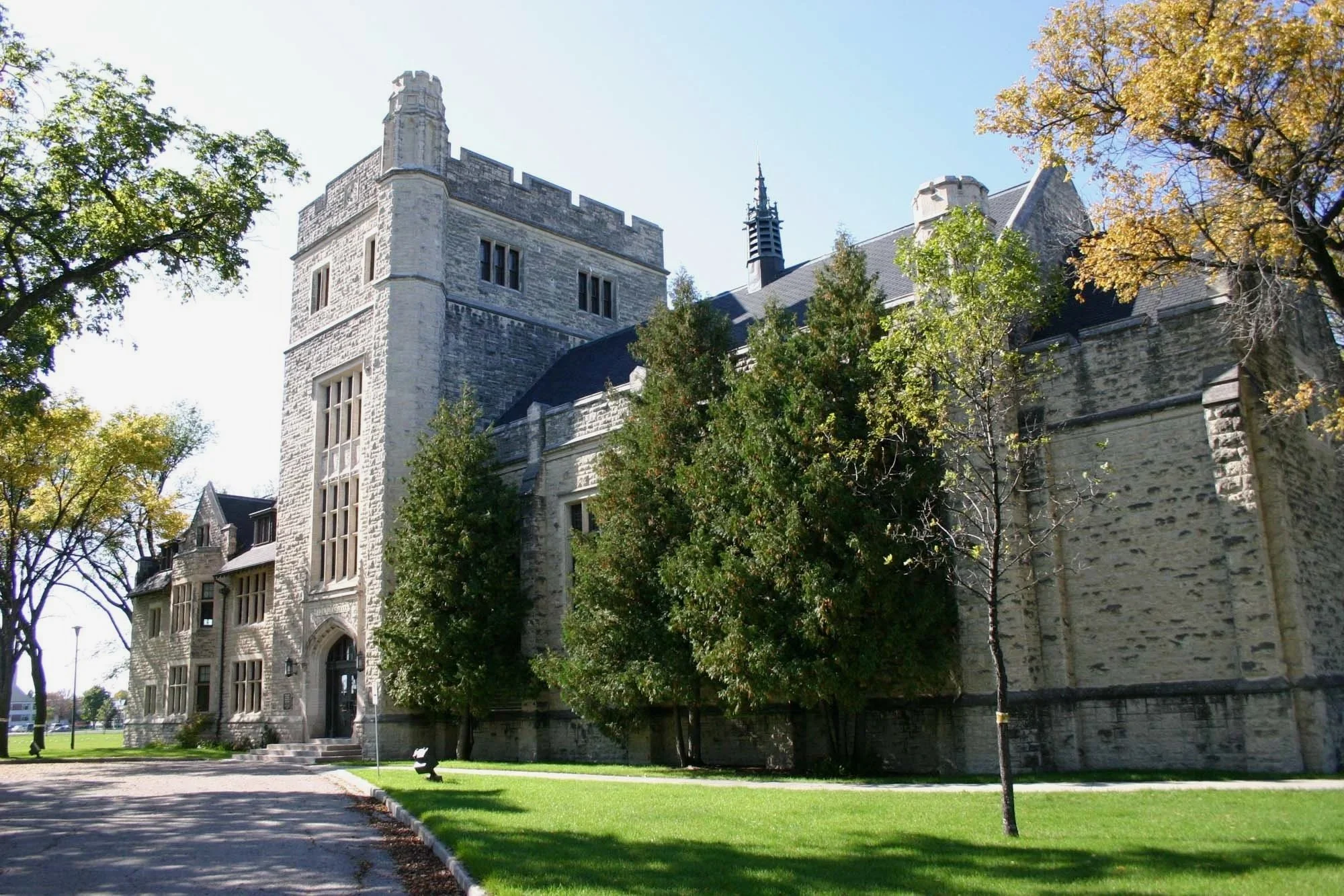 A historic stone castle with a tower, arched doorway, and tall windows, surrounded by trees and green grass on a sunny day.