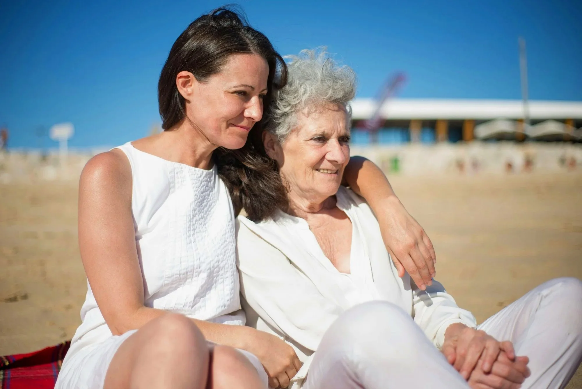 A young woman with long dark hair and an older woman with curly gray hair sitting together on a beach, smiling, with a blue sky and beach infrastructure in the background.