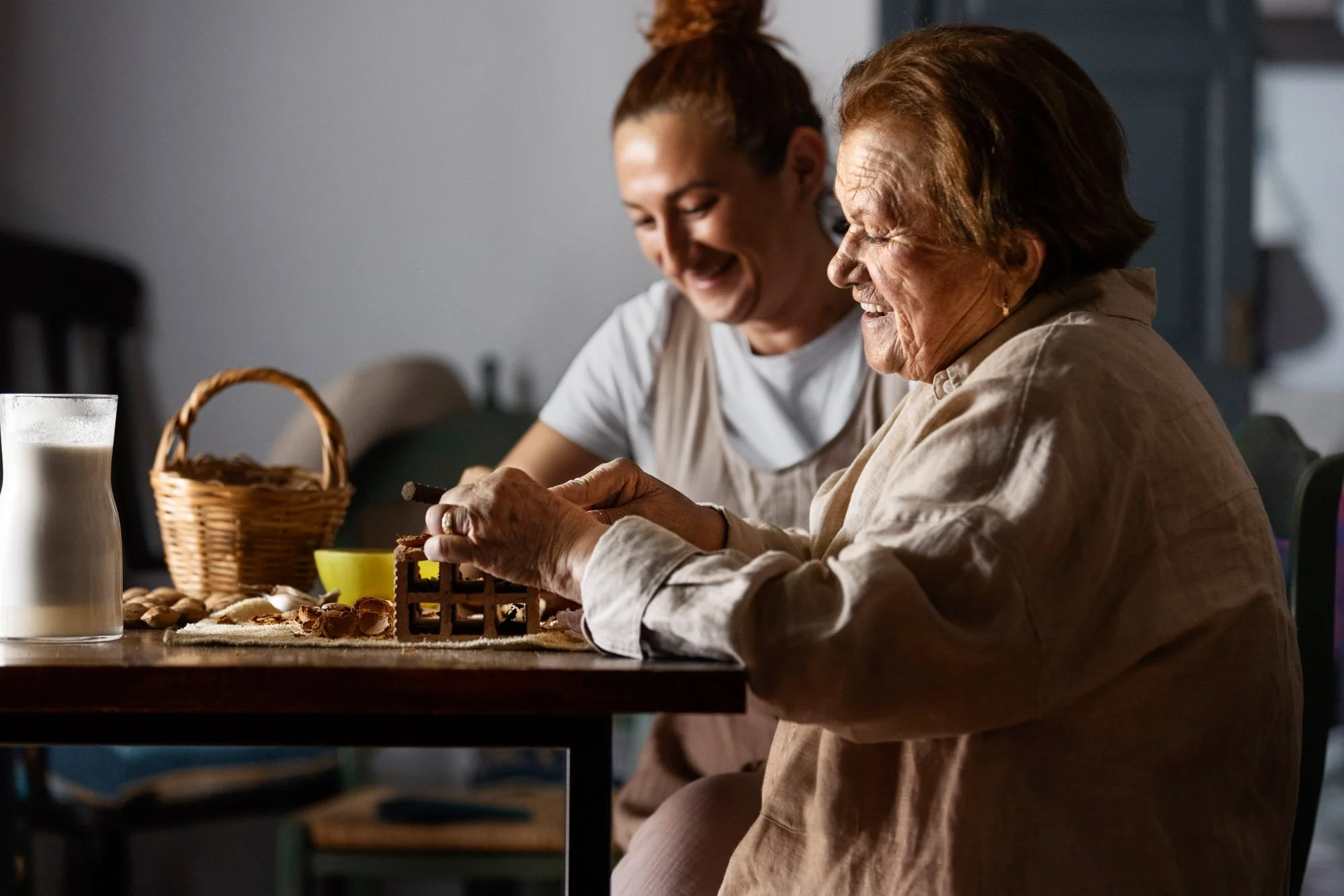 An elderly woman and a young woman enjoy each other's company at a table, cracking eggs and baking together. They are smiling and sharing a joyful moment.