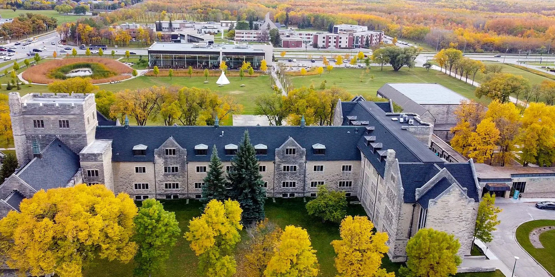An aerial view of a large historic building with a dark slate roof, surrounded by colorful autumn trees, on a university or campus area with parking lots and modern buildings in the background.