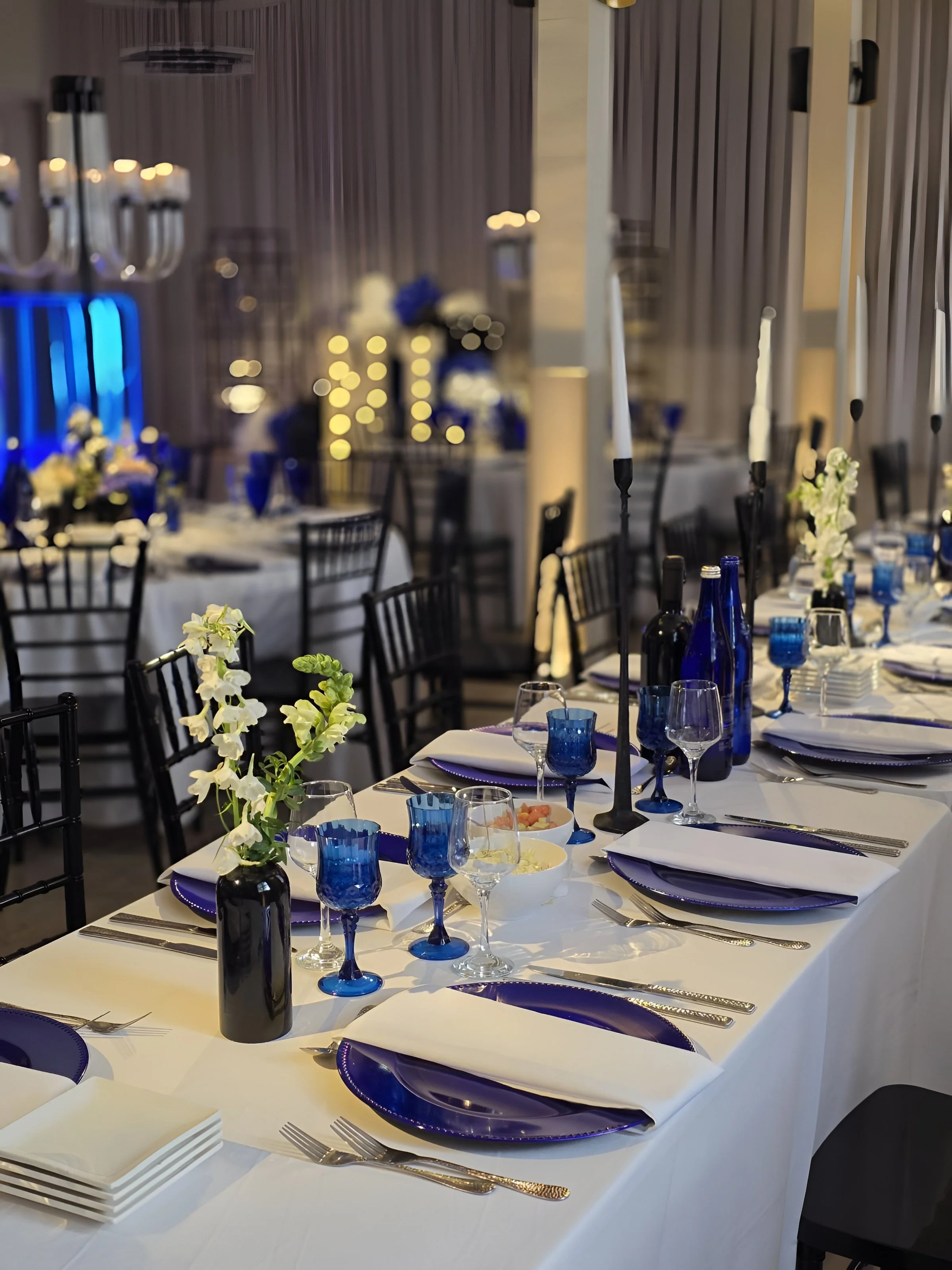 Table set for a formal event with white tablecloths, blue glassware, white and blue dinnerware, black chairs, tall candles, and floral centerpieces with white flowers and greenery.