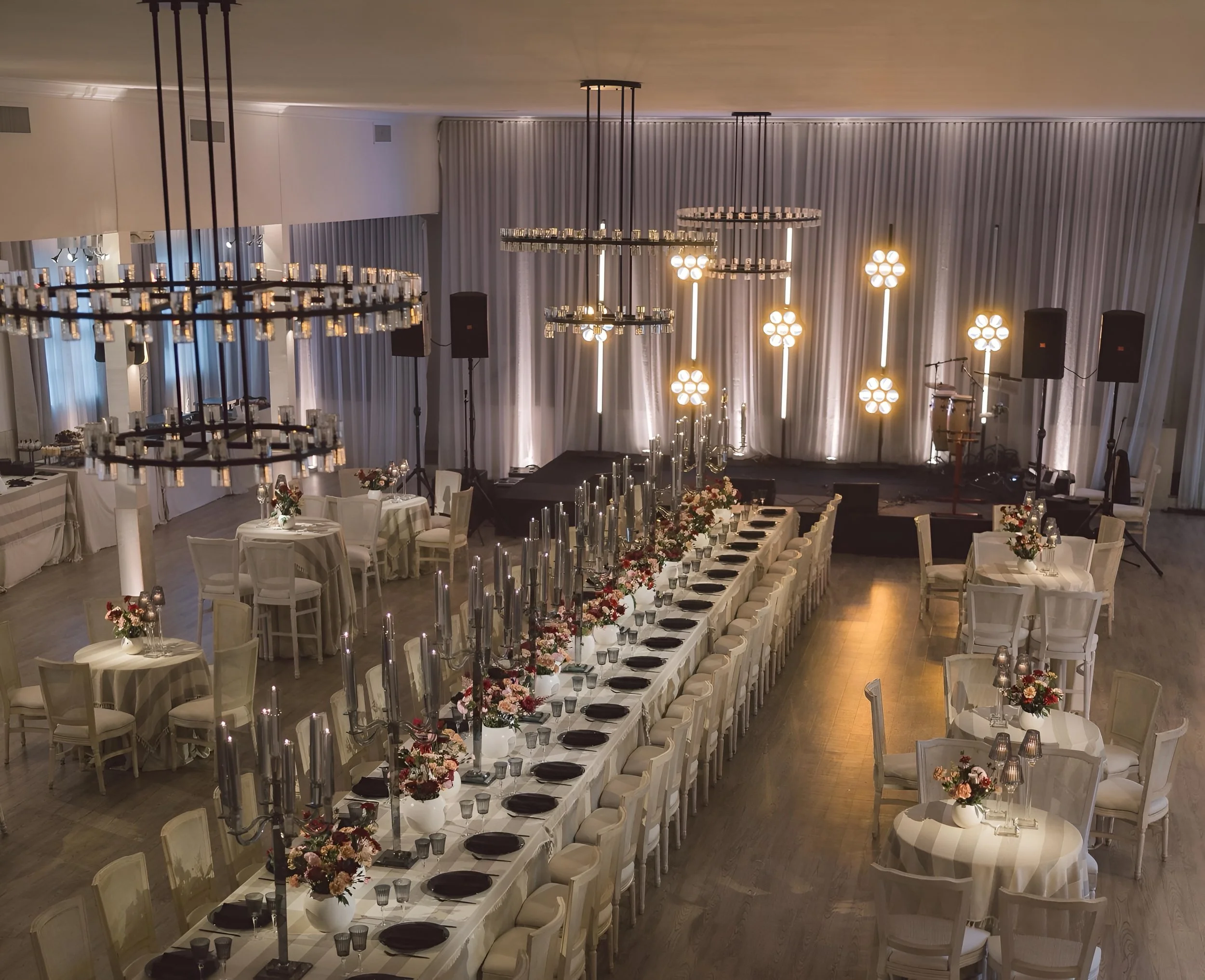 An elegantly decorated banquet hall with a long head table, floral centerpieces, candles, and round tables with flowers, set for a formal event, with a stage and lighting in the background.